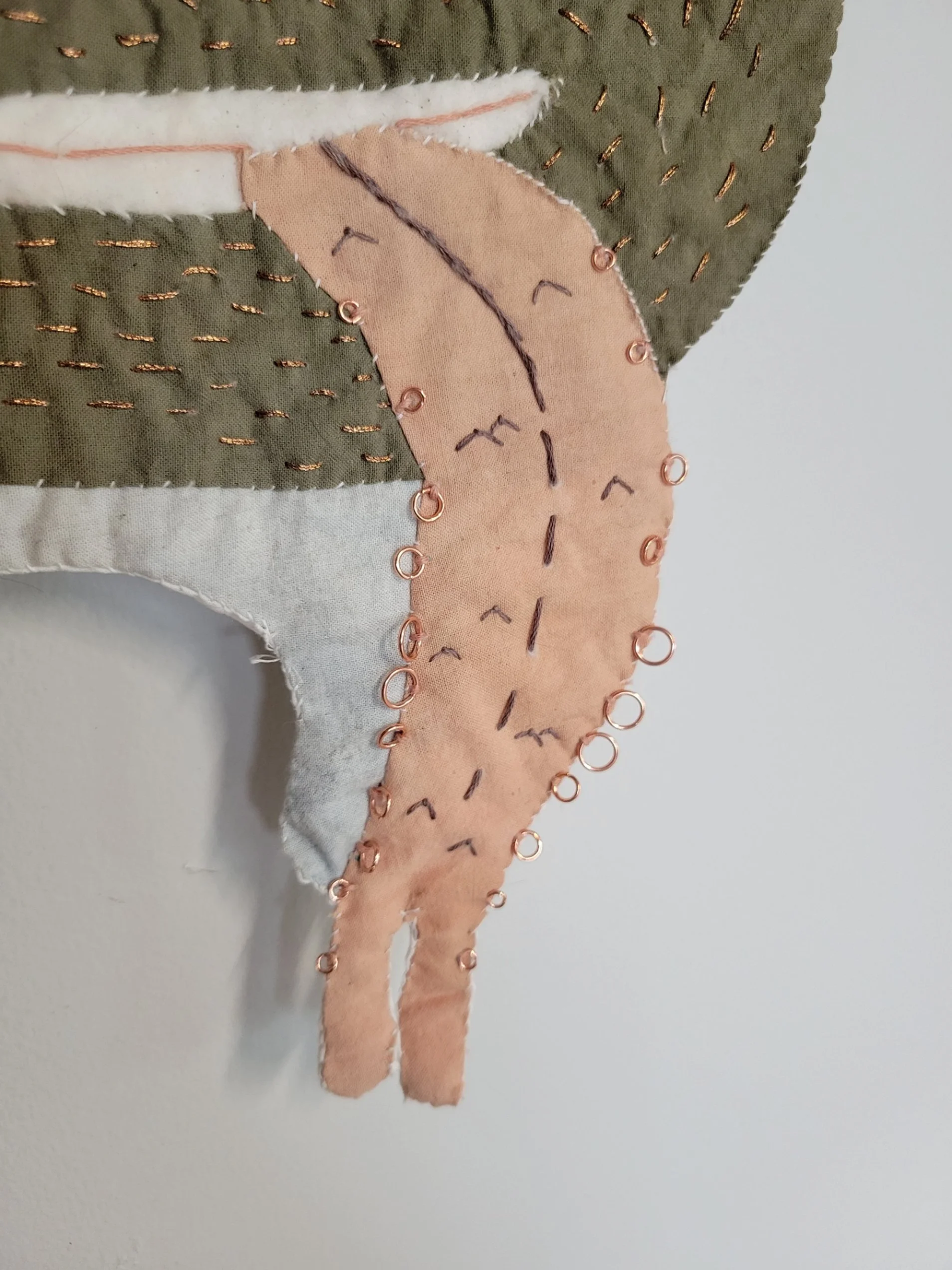 Close-up of a quilted fabric piece with stitched outlines, pins, and hand-stitched detail, featuring green, peach, white, and pink fabric sections.