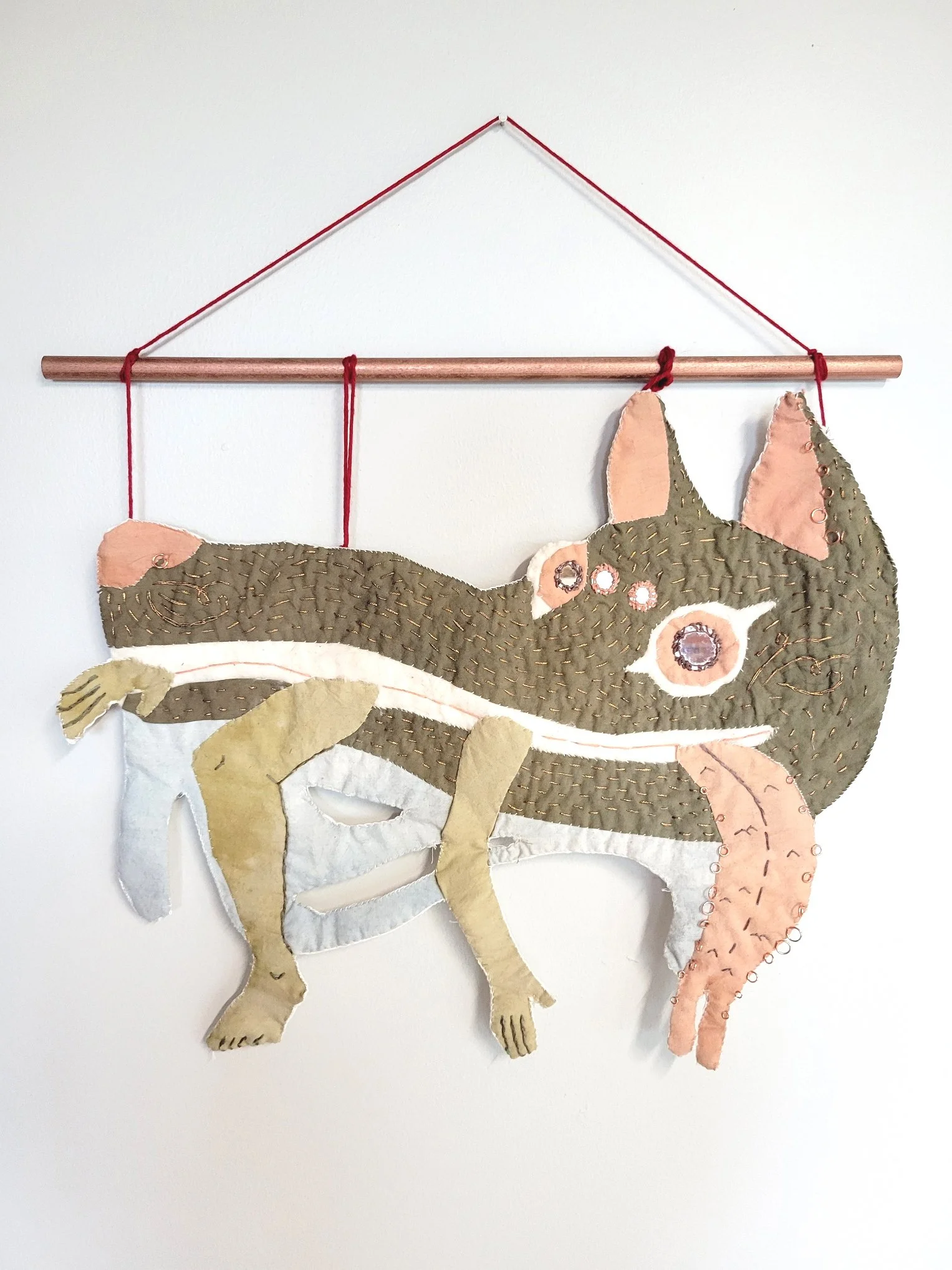 A fabric wall hanging of a chameleon with detailed stitching and patches, hanging from a copper rod attached to a red string on a white wall.