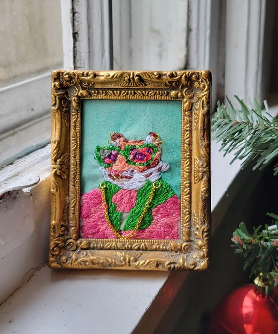  embroidered picture of a colorful leopard's head in a gold ornate frame, placed on a windowsill next to a decorated Christmas tree.