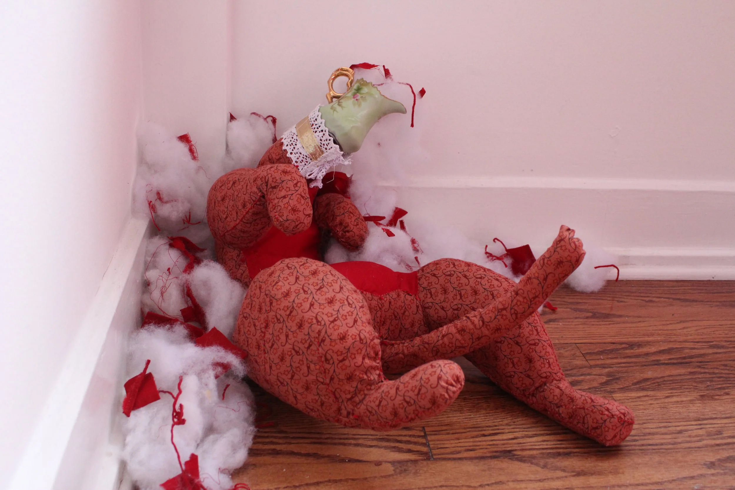 A broken stuffed fabric toy with a horse head, lying on a hardwood floor surrounded by cotton stuffing and red fabric scraps, in a corner of a room with pink walls.