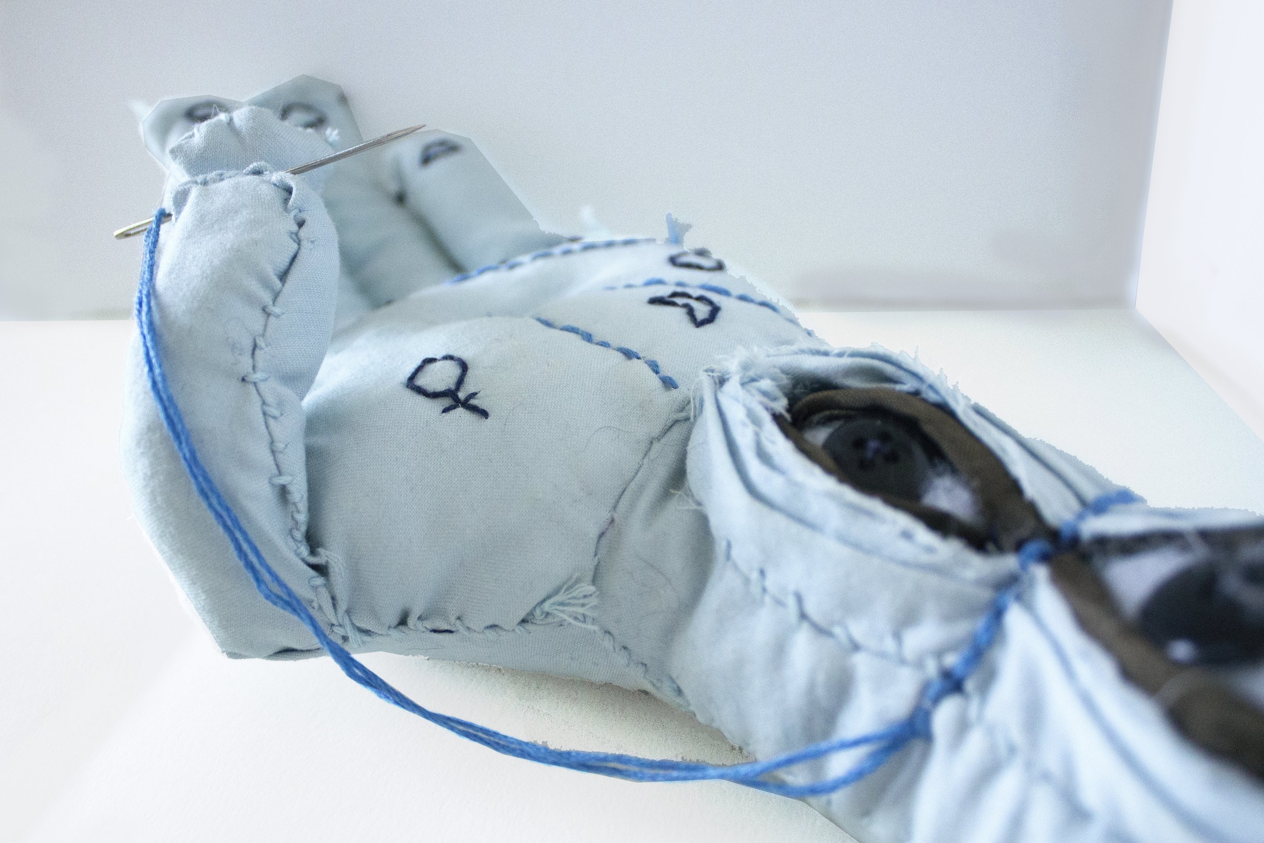 A fabric doll with embroidered facial features, dressed in a light blue outfit, with stitching details and buttons, lying on a white surface.