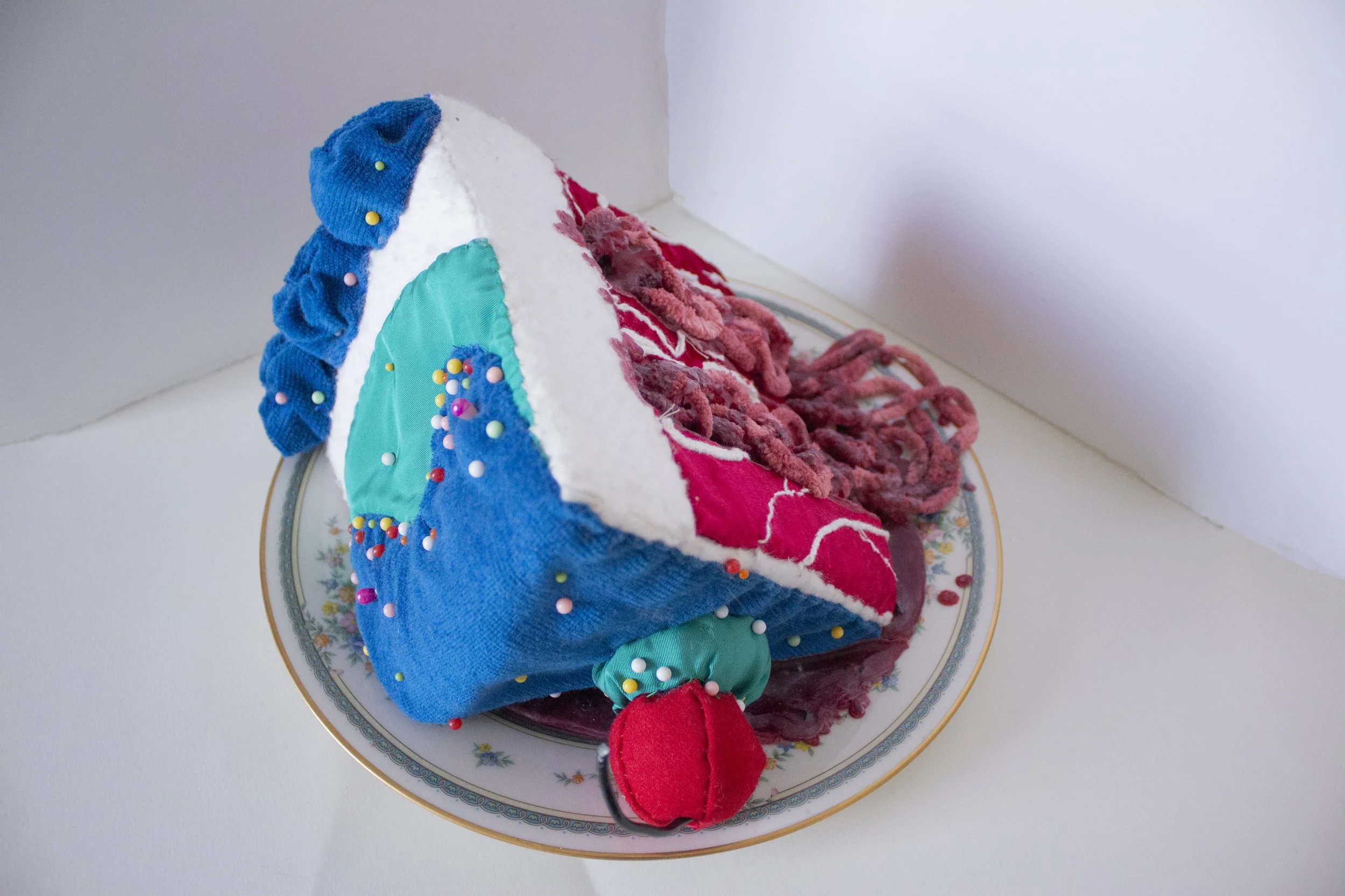 A colorful fabric art piece on a white plate with floral patterns. The artwork resembles a house with a blue roof, turquoise wall, white trim, and pink door, decorated with small colorful beads and pink pompom accents.