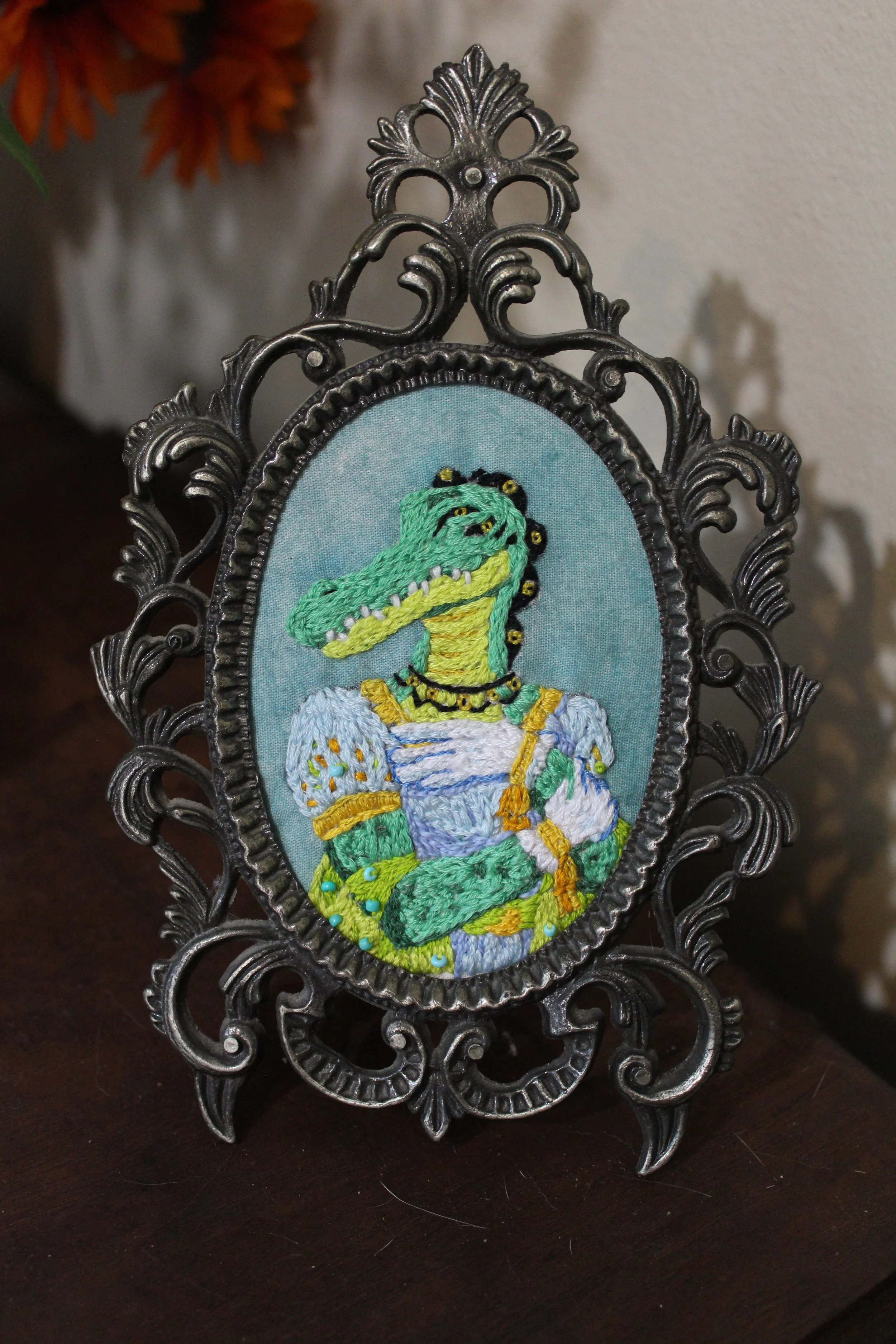 Embroidery of a cartoon crocodile dressed as a woman, wearing a blue dress with white puffs and gold accents, holding a parasol, in a decorative black metal frame.