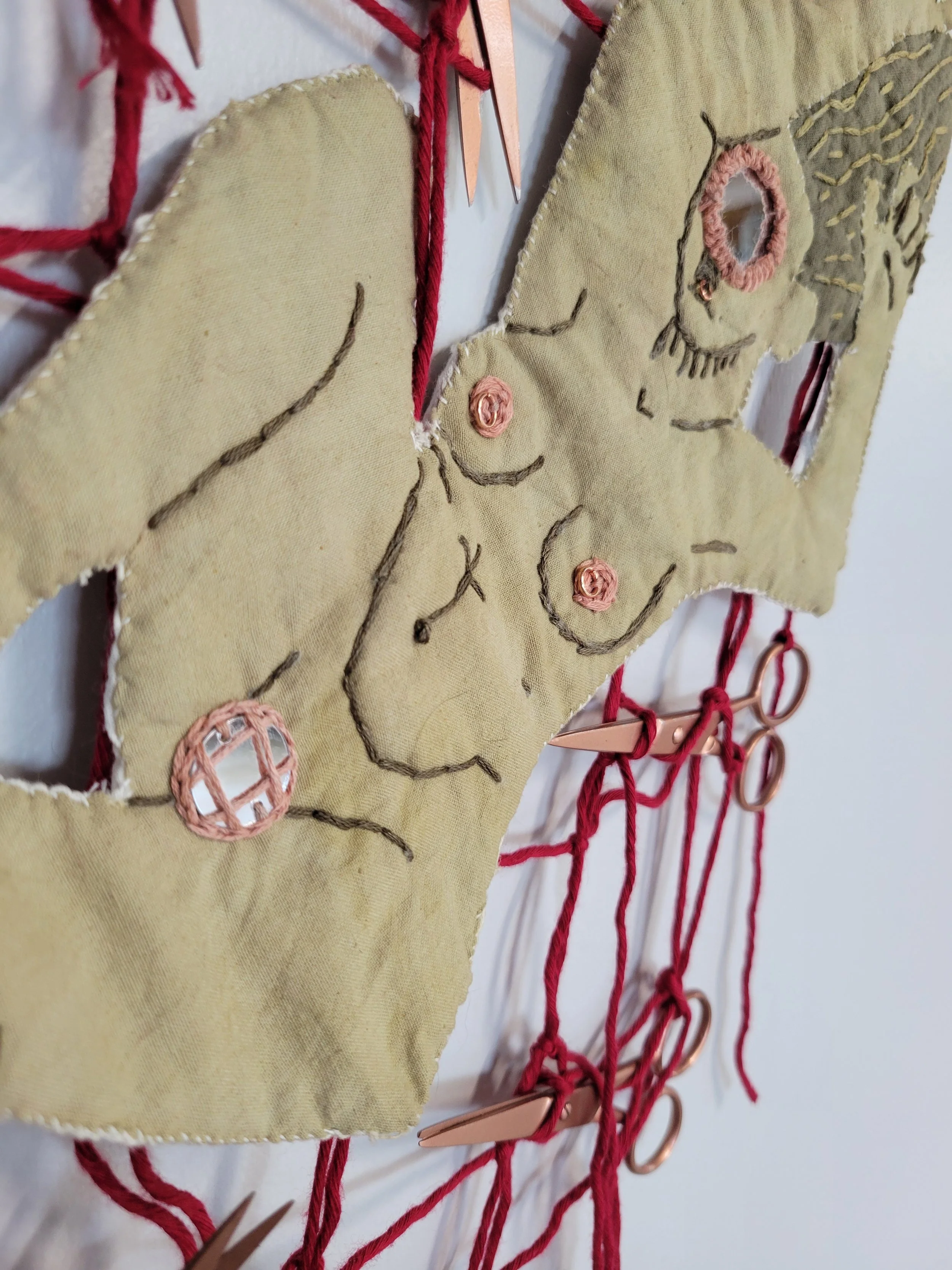 Close-up of a handmade craft depicting a nude female figure with embroidery details, mounted on a fabric background with red thread and metal rings.