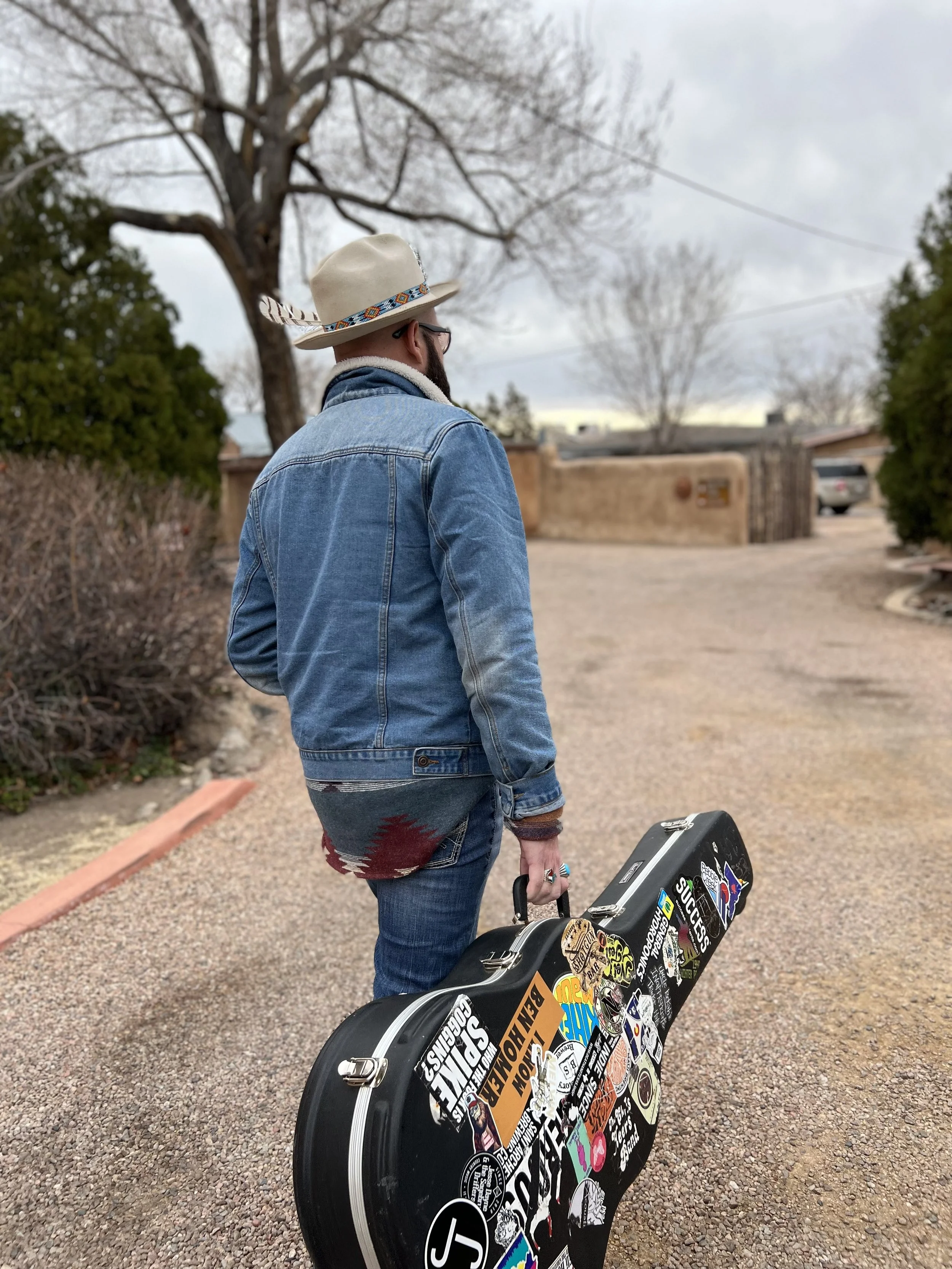 A man wearing a beige hat, glasses, a blue denim jacket, and a gray patterned shirt in a desert-like outdoor setting carrying a guitar case covered with various stickers.