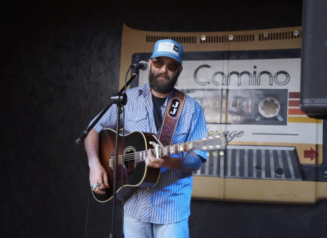 A man with a beard and sunglasses playing an acoustic guitar and singing into a microphone on stage, wearing a blue cap and striped shirt, with a retro cassette tape design in the background.