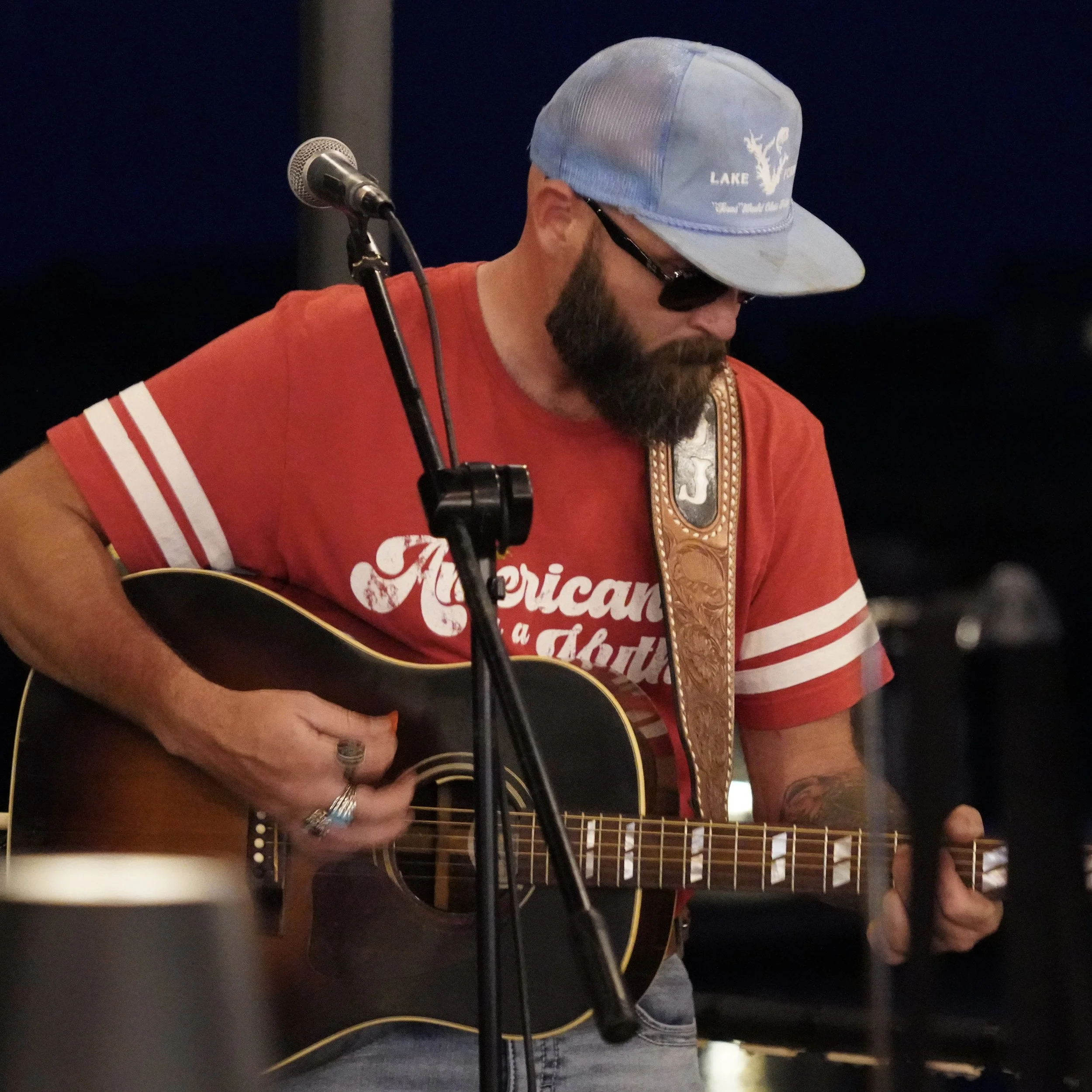 A man wearing a blue Cap, sunglasses, a red t-shirt, and playing an acoustic guitar.