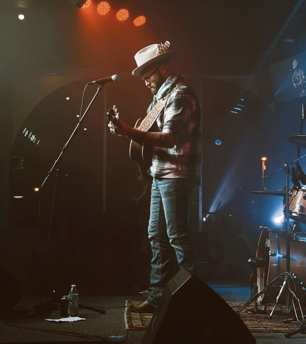 A man wearing a cowboy hat, sunglasses, and a plaid shirt is playing an acoustic guitar on stage under orange and blue lighting.