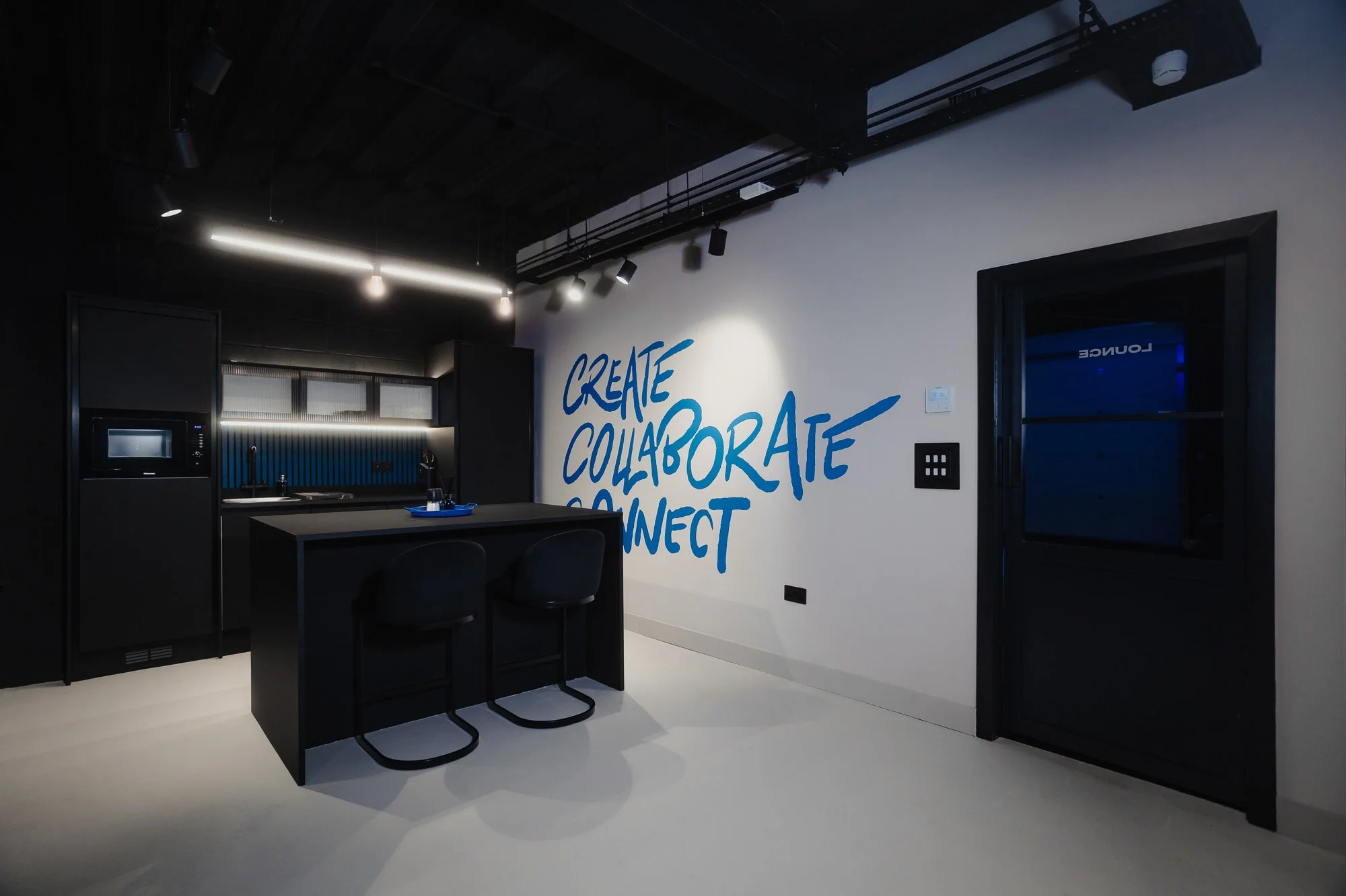 Modern kitchen with black cabinets and a white wall with blue text that says 'Create, Collaborate, Connect' in a stylish font, illuminated by track lighting.