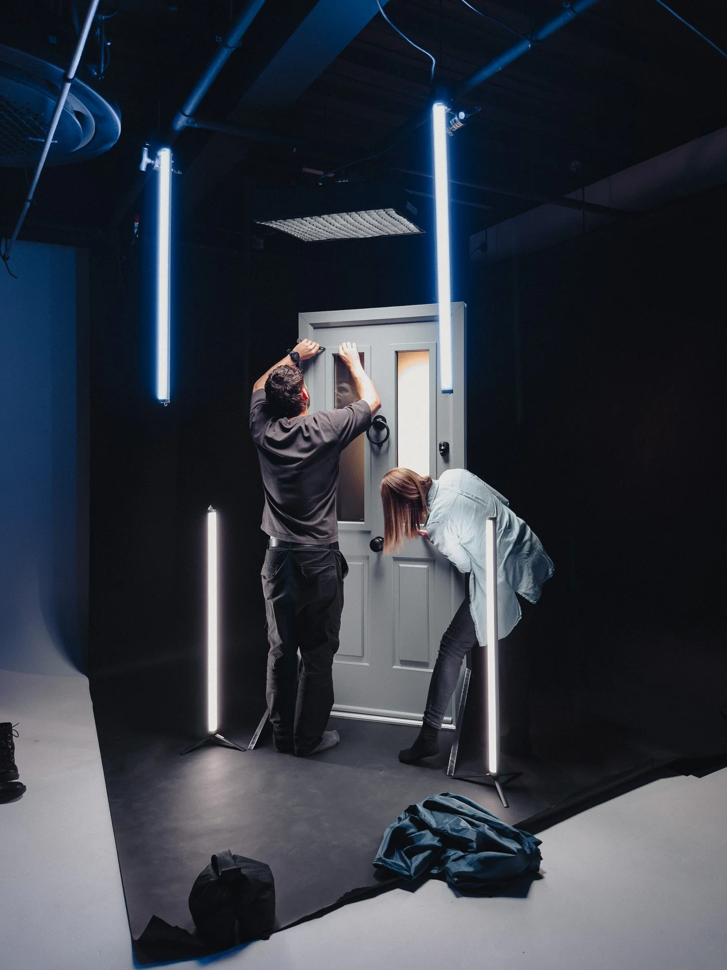 Two people setting up a door frame with a black backdrop, illuminated by vertical LED lights in a photography studio.