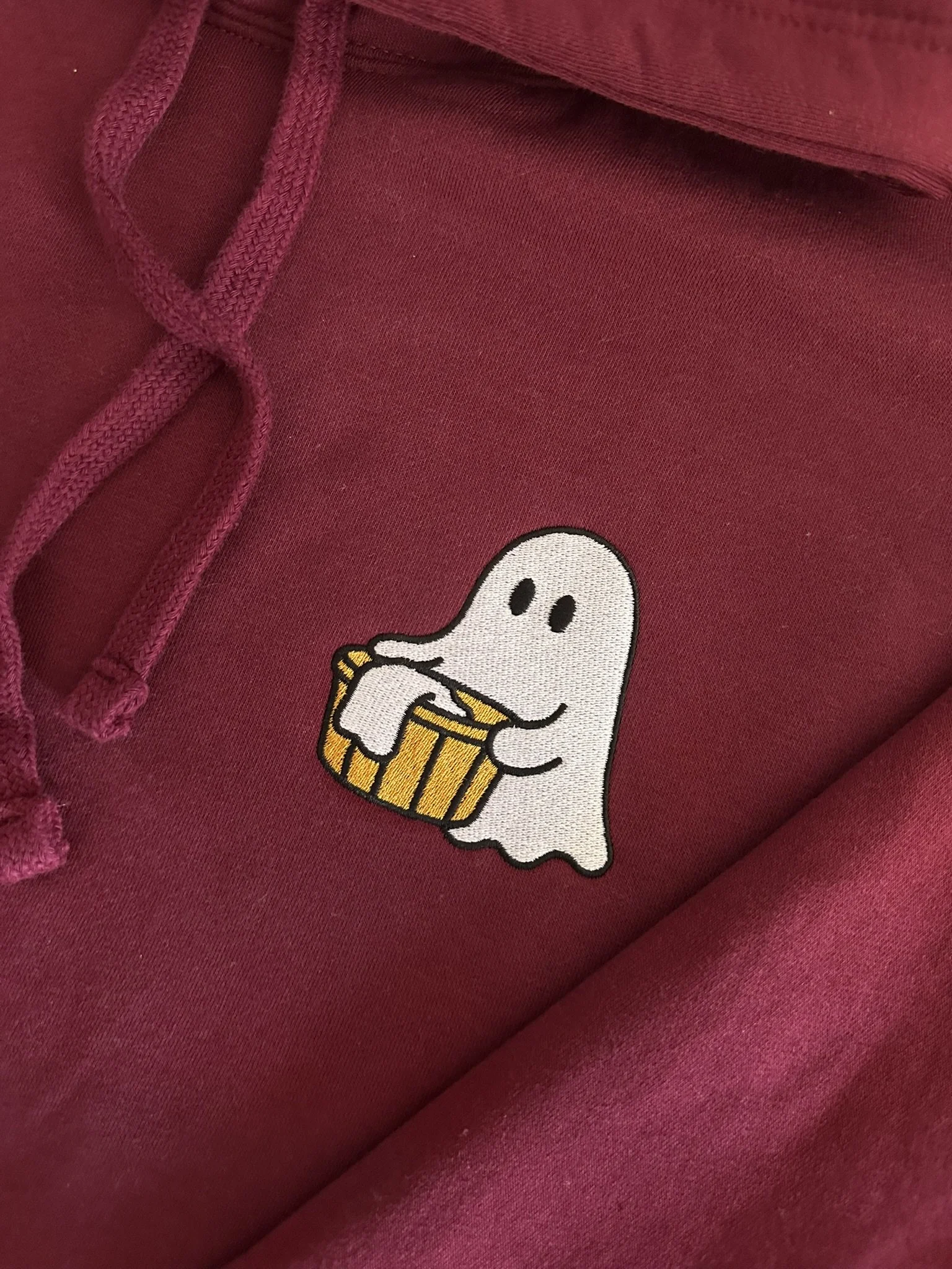 Close-up of ghost carrying a yellow laundry basket embroidered onto a burgundy hoodie