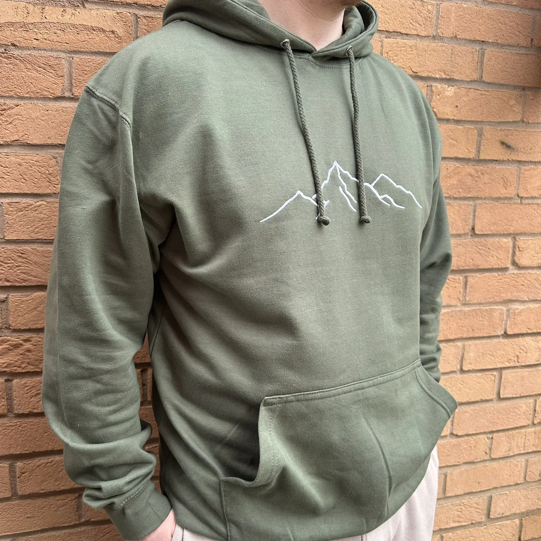 Model wearing a khaki hoodie with a minimalist white embroidered mountain design across the chest