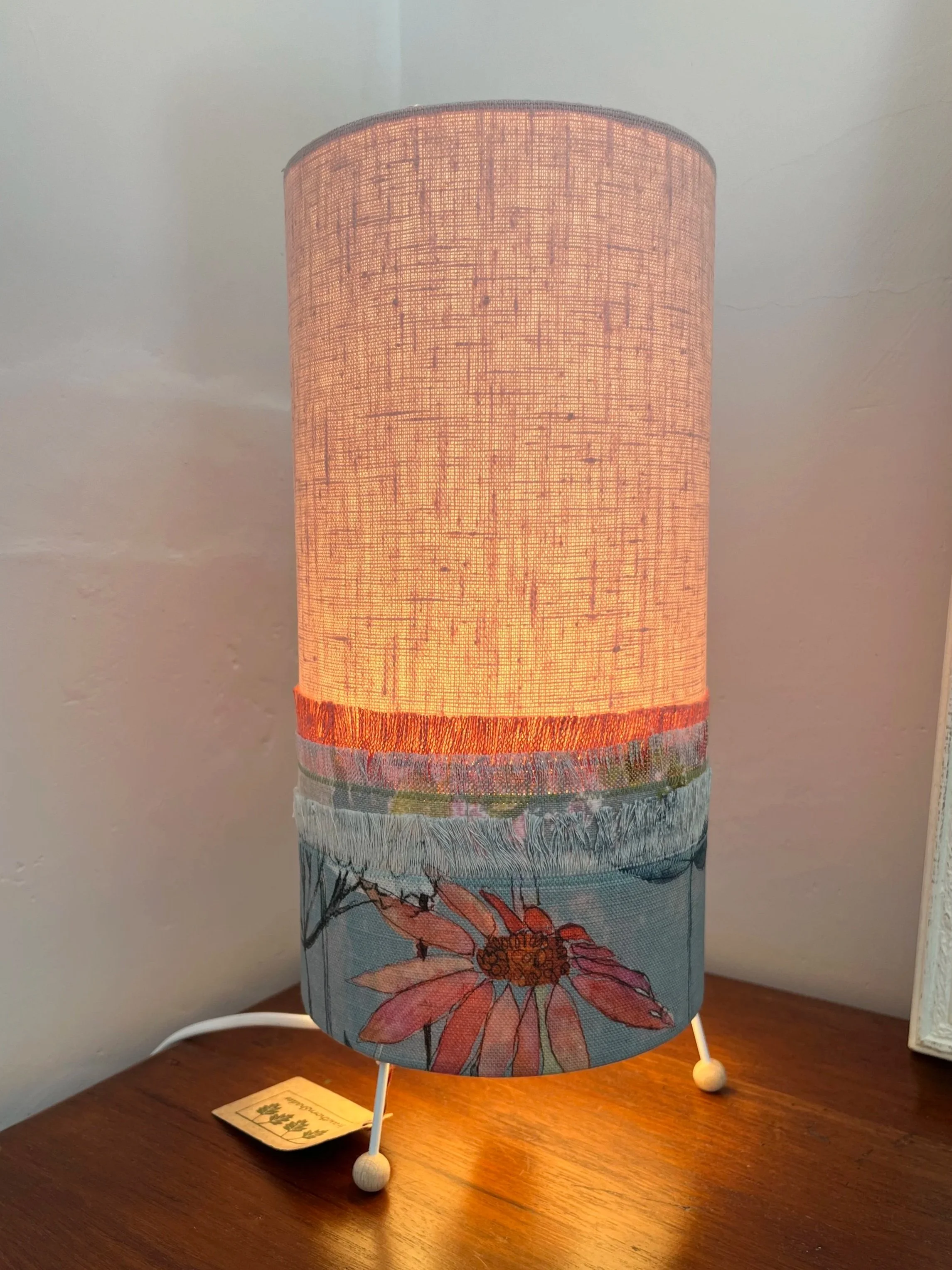 Cylindrical Table lamp with fringed fabric that resemble grasses, the shade is pale pink at the top, eith the lower section, pale blue with pink flowers.