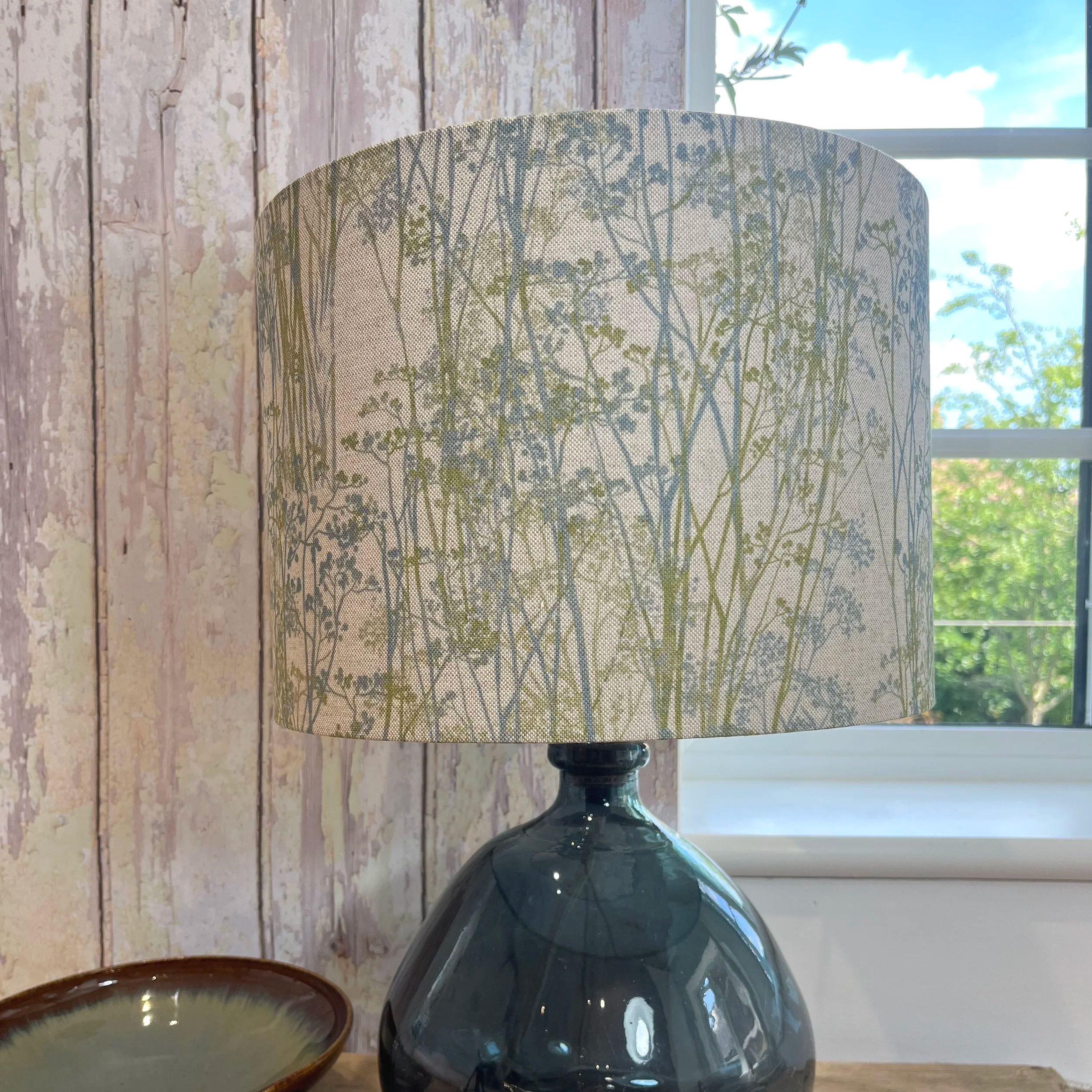 Hedgerow grass printed drum shape lampshade in blue grey and green on natural linen sitting on a teal blue glass lamp base.