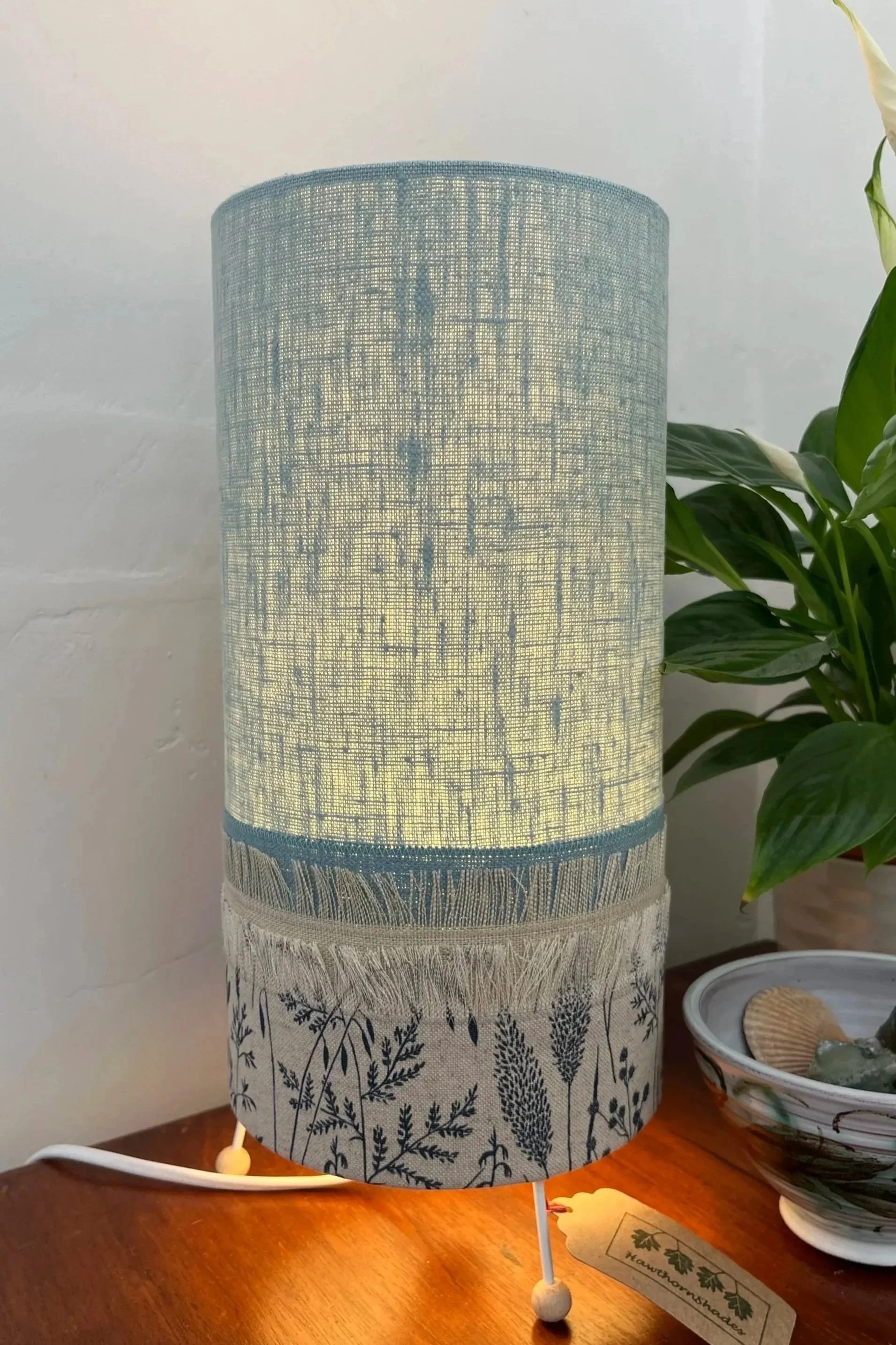 Cylindrical table lamp made with fringed and appliqued fabrics, top section pale blue, lower section is cream with dark blue grasses.
