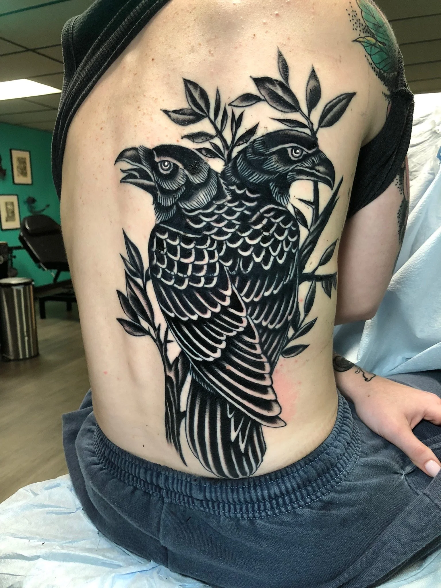 Black tattoo of a hawk with detailed feathers and leaves on person's side torso.
