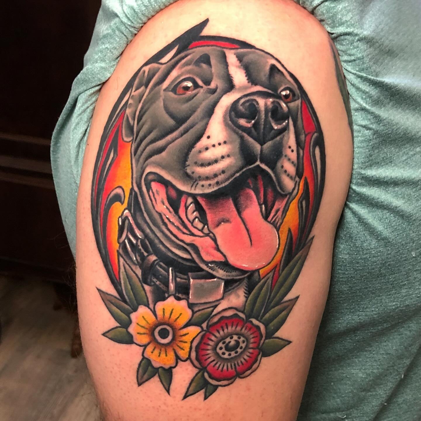 Colorful tattoo of a dog's face, possibly a pitbull or similar breed, with its tongue out, surrounded by flames and flowers on upper arm.
