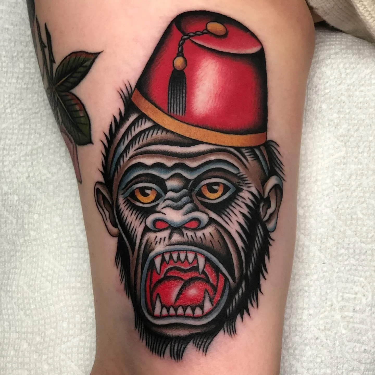 Tattoo of a gorilla's face with an open mouth showing sharp teeth, wearing a red hat with a tassel, on a person's thigh.