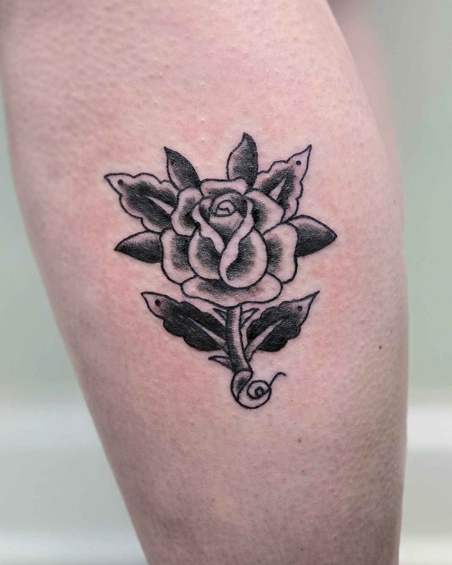 Black and gray rose tattoo on skin featuring a detailed rose with leaves and a swirling stem.