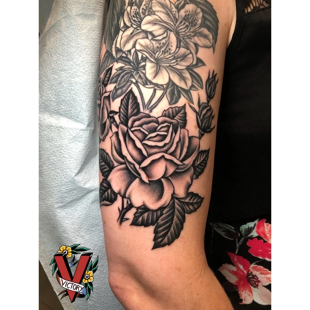 A detailed black and gray floral tattoo on a person's upper arm featuring a large rose and other flowers with leaves.