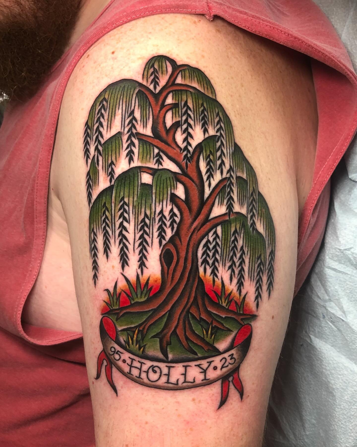A colorful tattoo of a weeping willow tree with green leaves and brown branches on a person's upper arm. A banner beneath the tree reads '95·HOLY·23'.