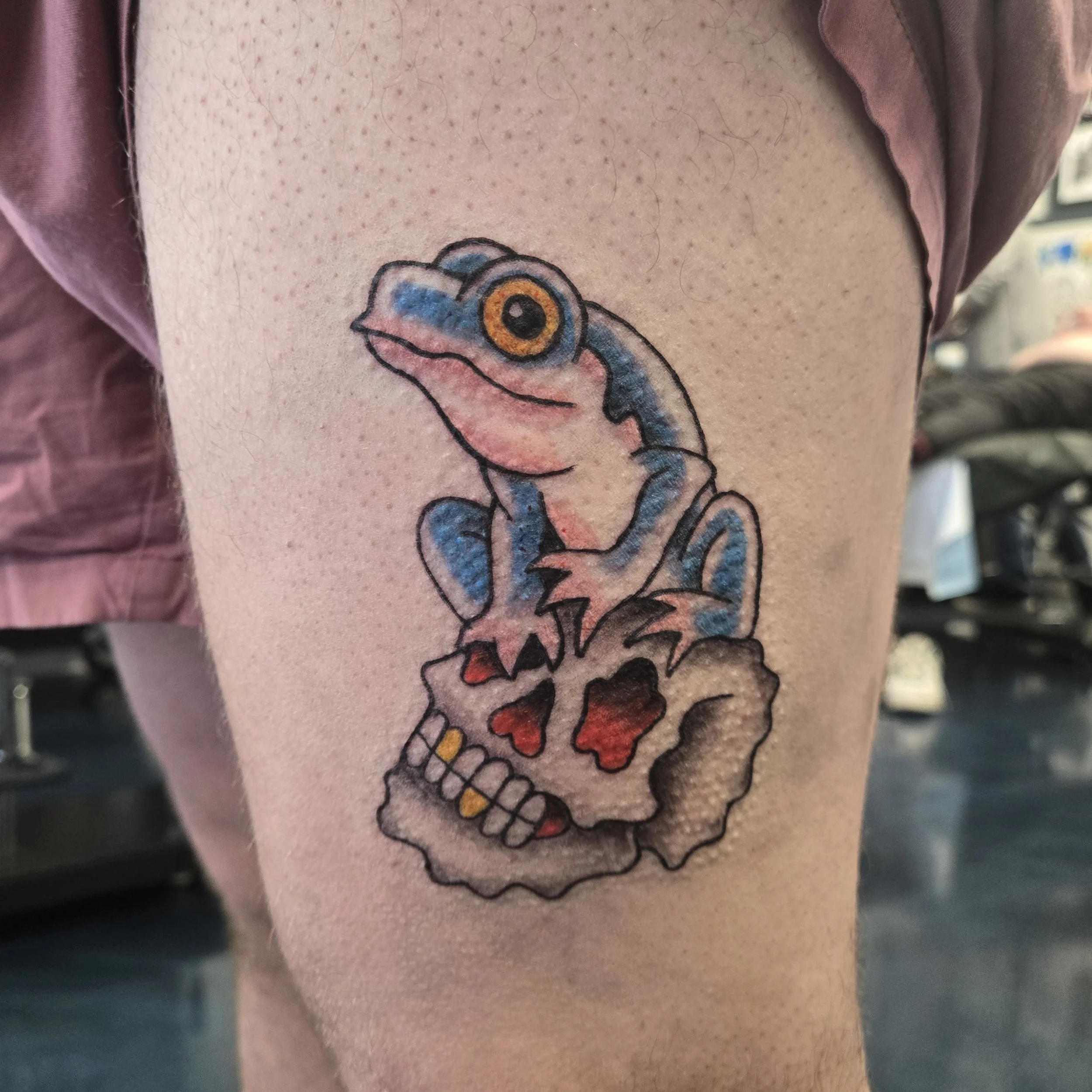 Colorful tattoo of a blue gecko with large orange eyes sitting on a skull with red eyes and a wide grin.