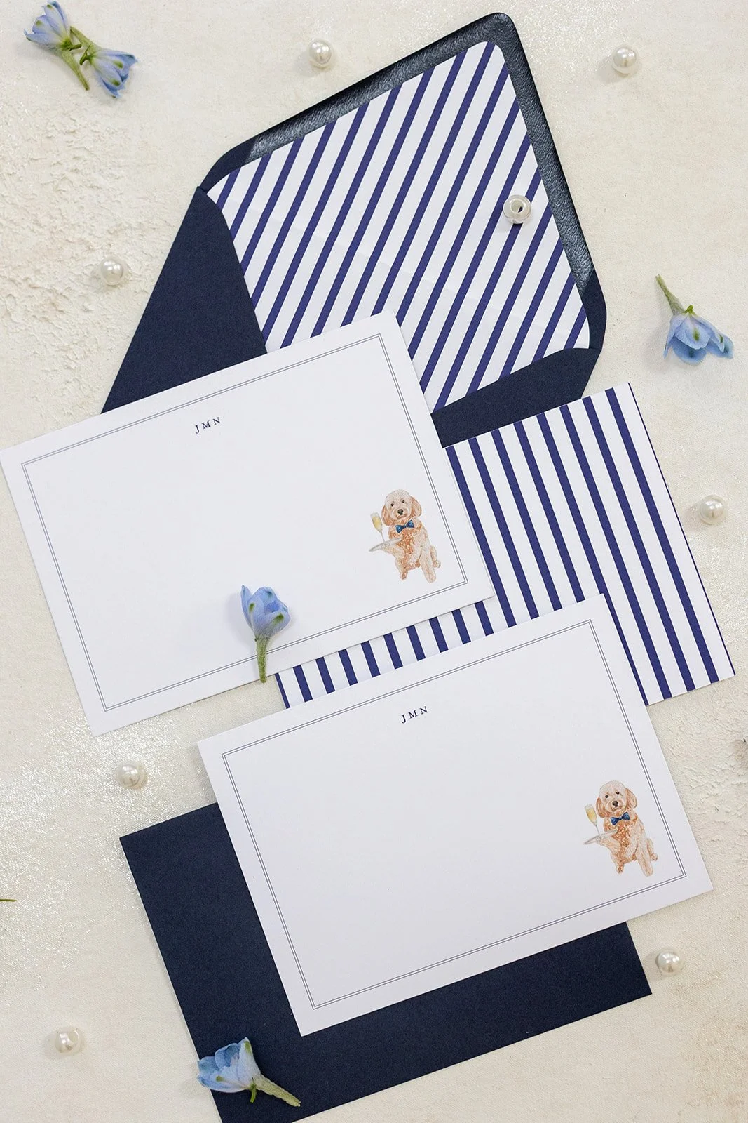 Two white cards with a dog illustration, orange and fluffy, wearing a blue bow tie, placed on top of navy blue and white striped envelopes, with small blue flowers and pearl decorations scattered on a white textured background.