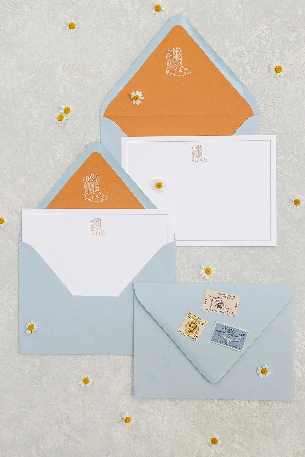 Three light blue envelopes with white and orange letter papers featuring illustrations of cowboy boots and a stamp with a cowboy riding a bucking horse. Small daisies are scattered around on a light textured surface.