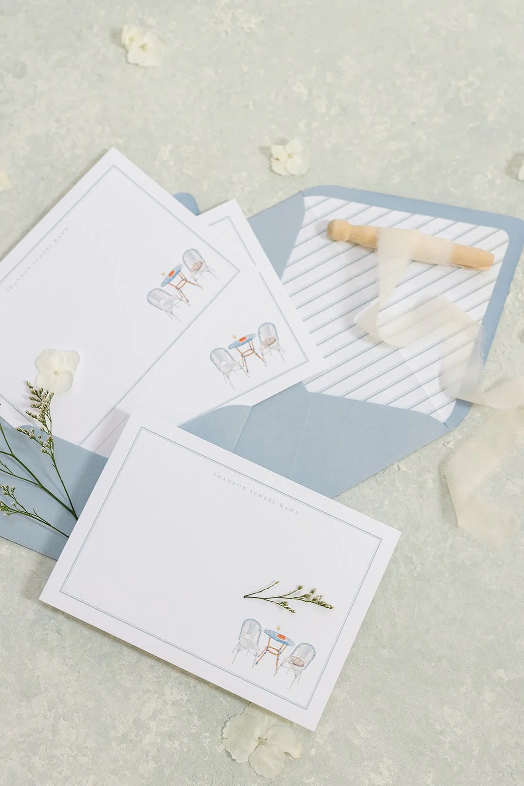 Wedding invitation suite with light blue envelope, floral illustrations, and a notepad with a band-aid on it, laid on a textured surface with scattered flowers.