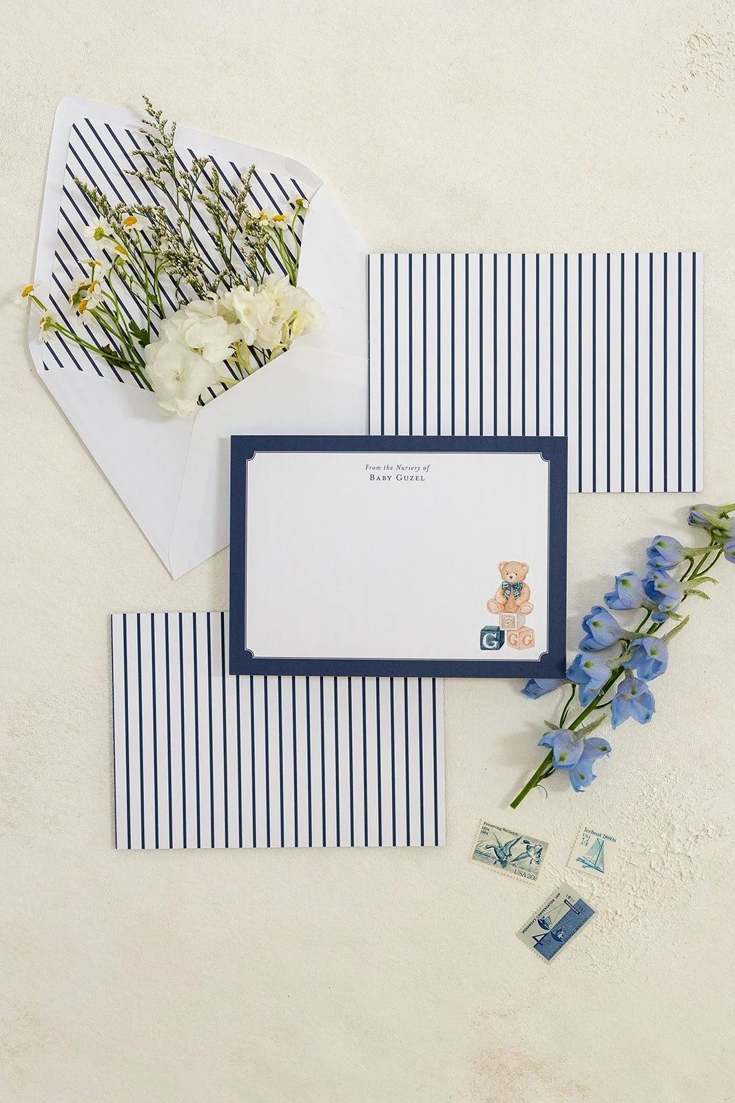 Baby shower invitation with blue striped paper, a small teddy bear illustration, and the text 'From the Nursery of Baby Guzel'. Includes a floral arrangement of white and blue flowers, and vintage postage stamps.
