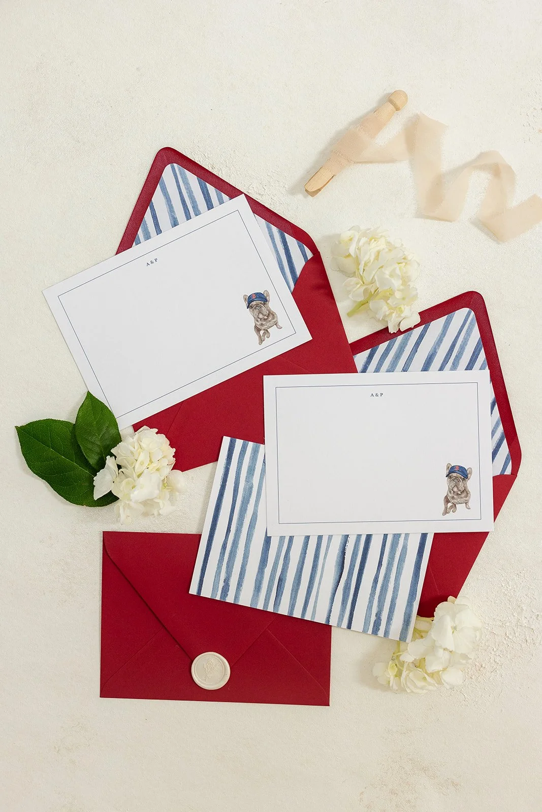 Red envelopes containing with striped blue and white interior lining, and illustrated custom stationary cards with art of a bulldog wearing a blue cap, surrounded by white flowers, green leaves, and a small wooden clothespin with a ribbon.