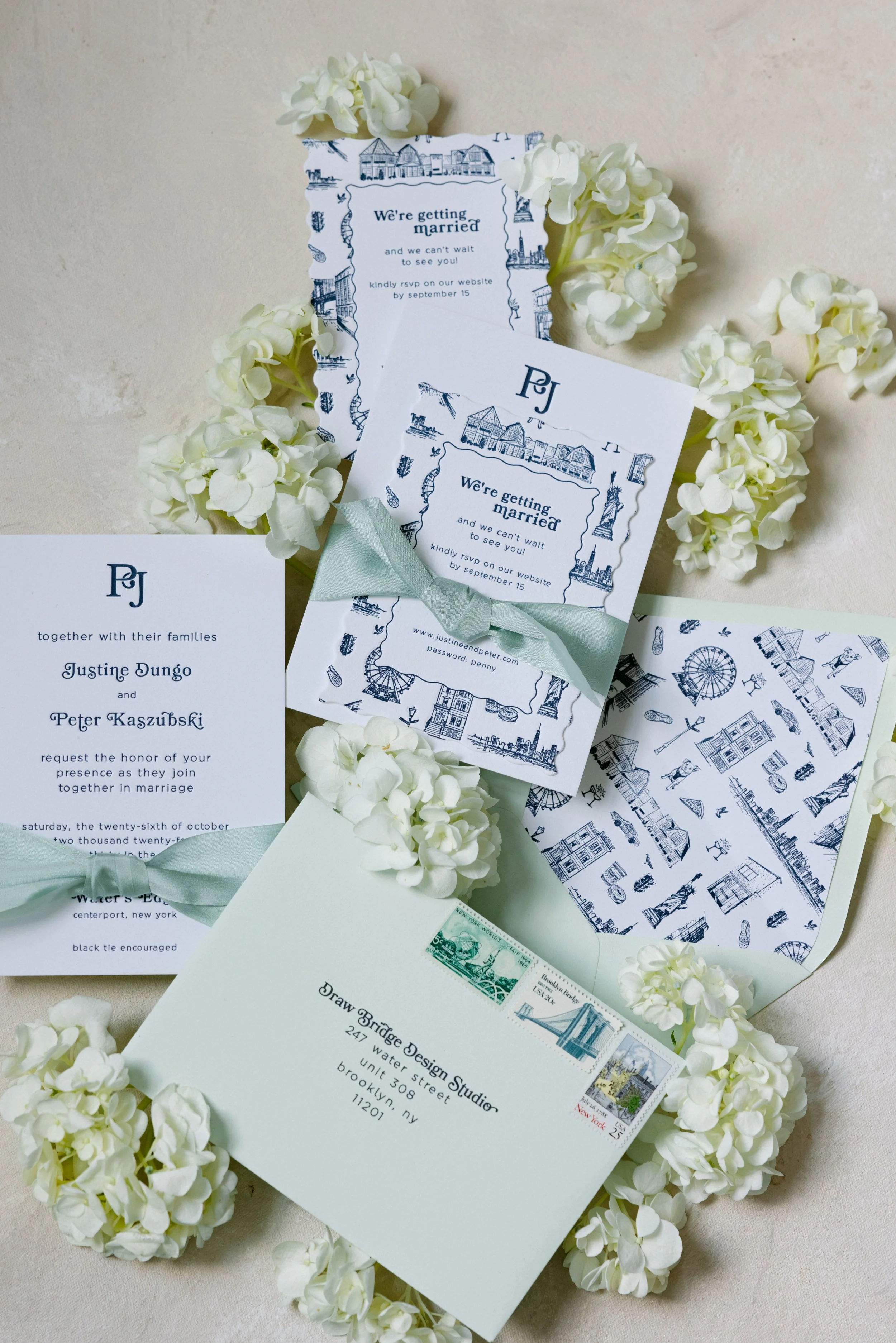 A custom wedding invitation suite arranged with white hydrangea flowers, including custom ribbon elements, custom invitation envelope interior lining artwork, vintage US postage stamps, an invitation card, a details card, an RSVP card, and custom art