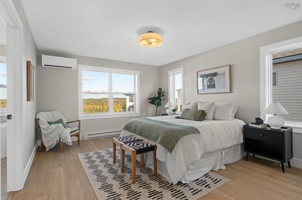A bedroom with a large bed covered in white and beige bedding, a green throw blanket, pillows, and a rug underneath. There are two bedside tables with lamps, a window, a chair with a blanket, and a framed picture on the wall.
