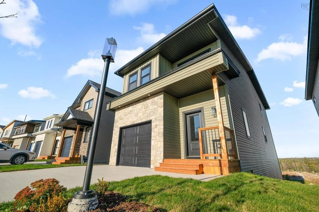 Modern residential house with a small front porch, attached garage, green siding, and stone accents, located in a neighborhood with similar houses under a partly cloudy sky.