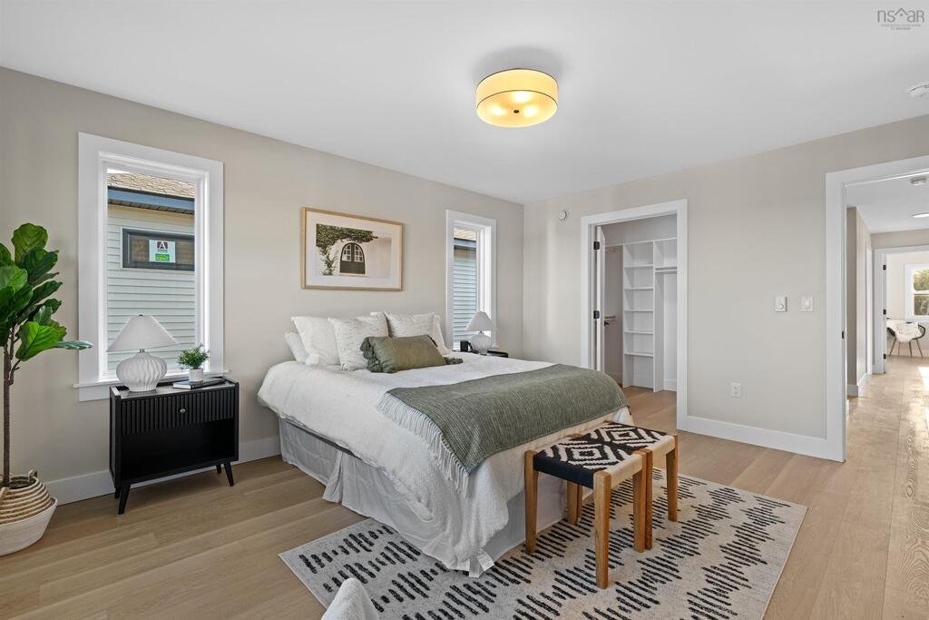 A bedroom with a white bed, decorative pillows, and a gray blanket. Nightstands with lamps and small plants. Light wood flooring, a patterned rug, and two windows. An open closet and a hallway leading to another room.