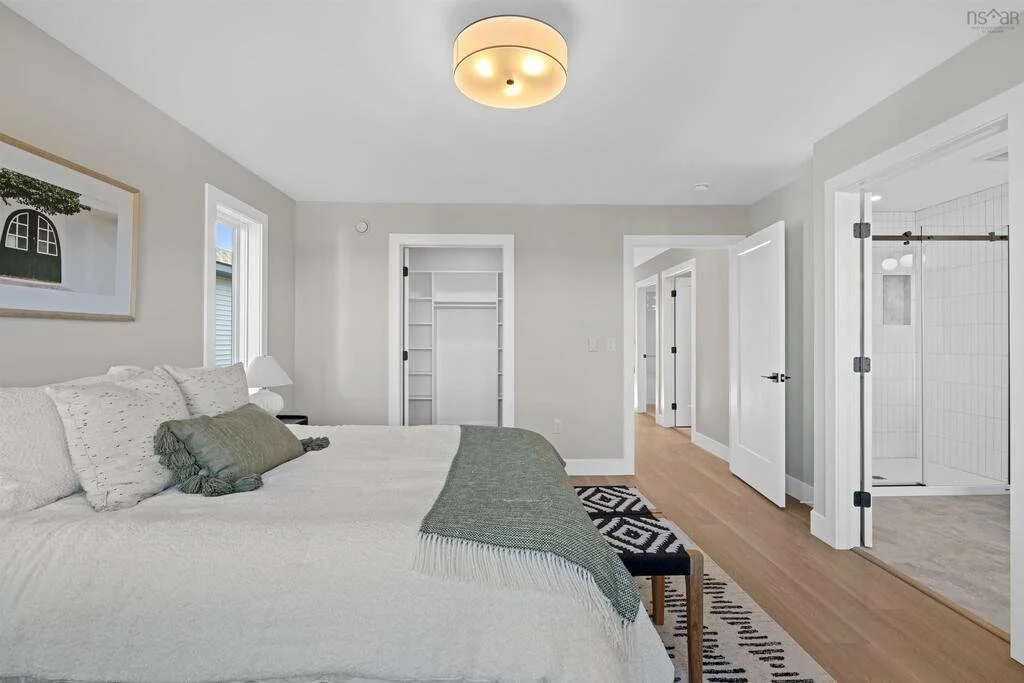 A bedroom with a bed, nightstand, lamp, and window. The room has light-colored walls, a wooden floor, and an open doorway leading to a bathroom with a walk-in shower.