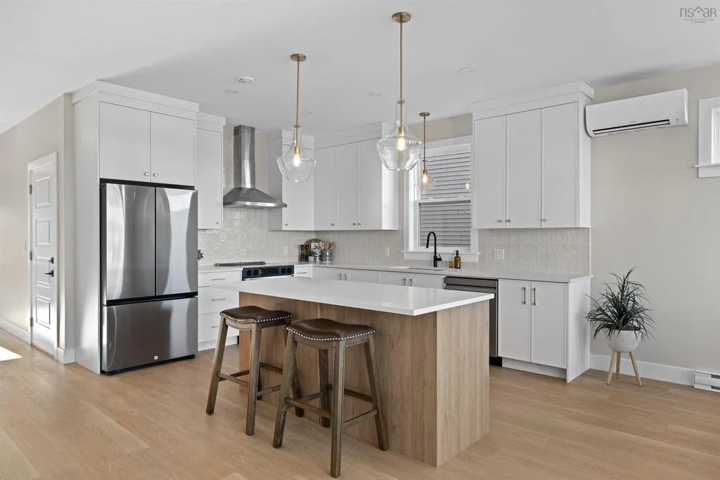 Modern kitchen with white cabinets, stainless steel refrigerator, island with wooden base and two bar stools, stainless steel range hood, white subway tile backsplash, and light wood flooring.