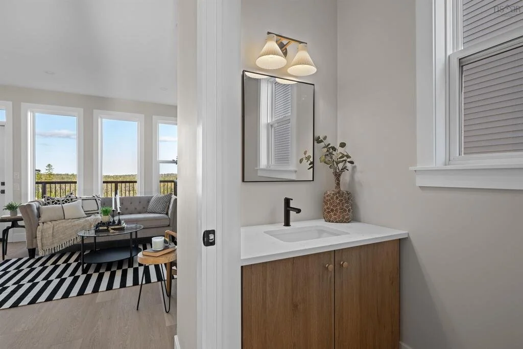 A small bathroom vanity with a white countertop and a black faucet, a mirror above, a small window, and a decorative plant in a textured vase. The bathroom is adjacent to a bright living room with large windows, a striped black and white rug, and sea