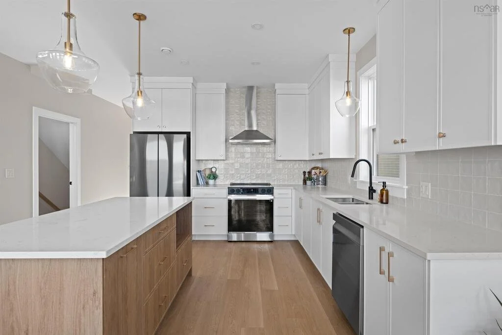 Modern white kitchen with wooden accents, stainless steel refrigerator, oven, and dishwasher, pendant lights, and a window above the sink.