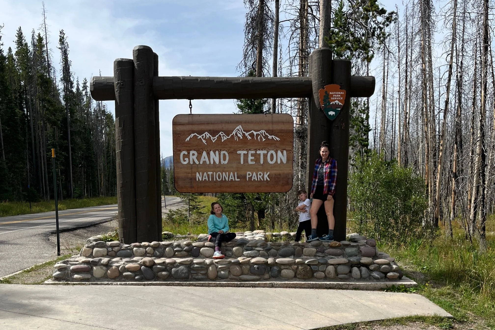 The author and her kids standing in front of the Grand Teton National Park entrance sign