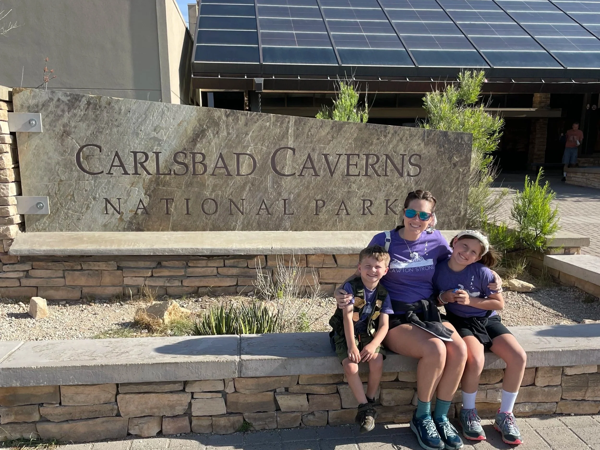The author and her children sitting in front of the visitors center and large sign for Carlsbad Caverns