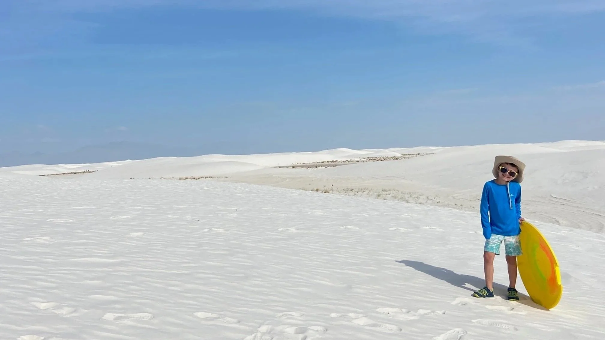 Boy in bright blue sunshirt and khaki hat holds a yellow sand sled in the dune field of White Sands National Park