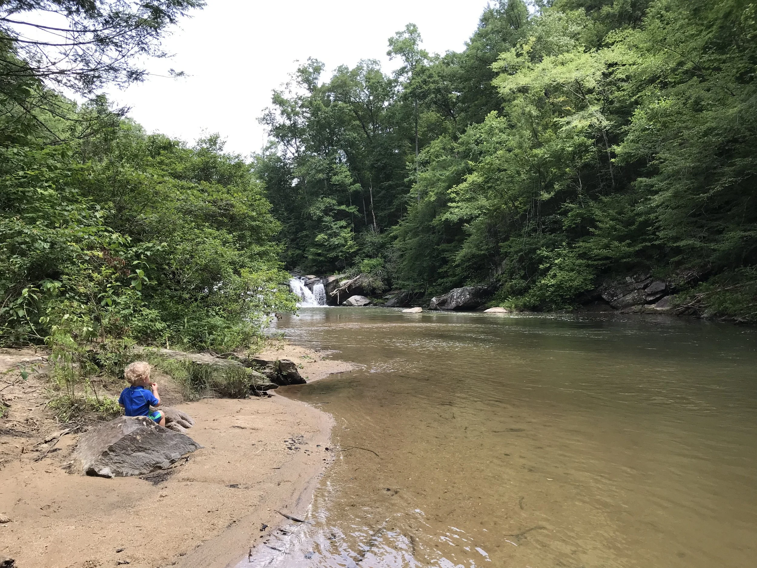 The author's toddler son sits on a rock and eats a snack while watching a waterfall