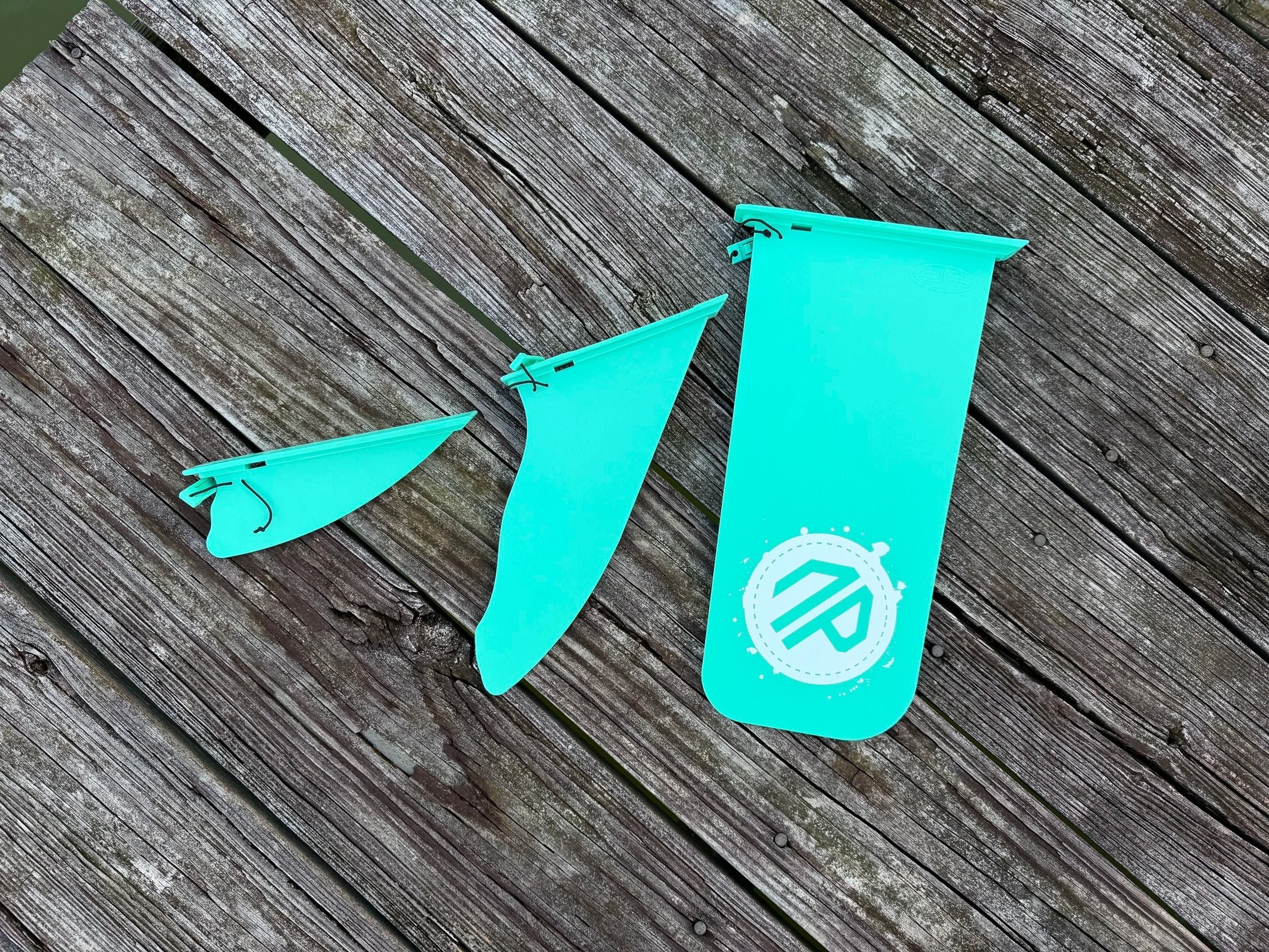 3 aqua colored paddle board fins are placed in a flat lay on a wooden dock