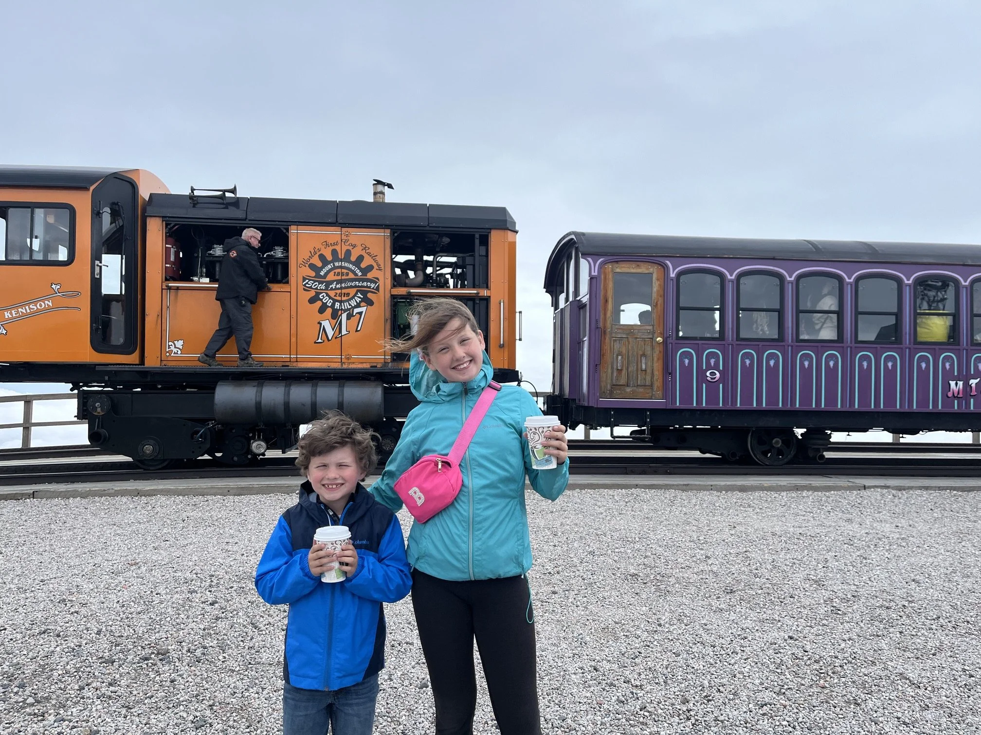 The author's children in colorful rain coats holding hot cocoa cups and standing in front of the trains at the summit