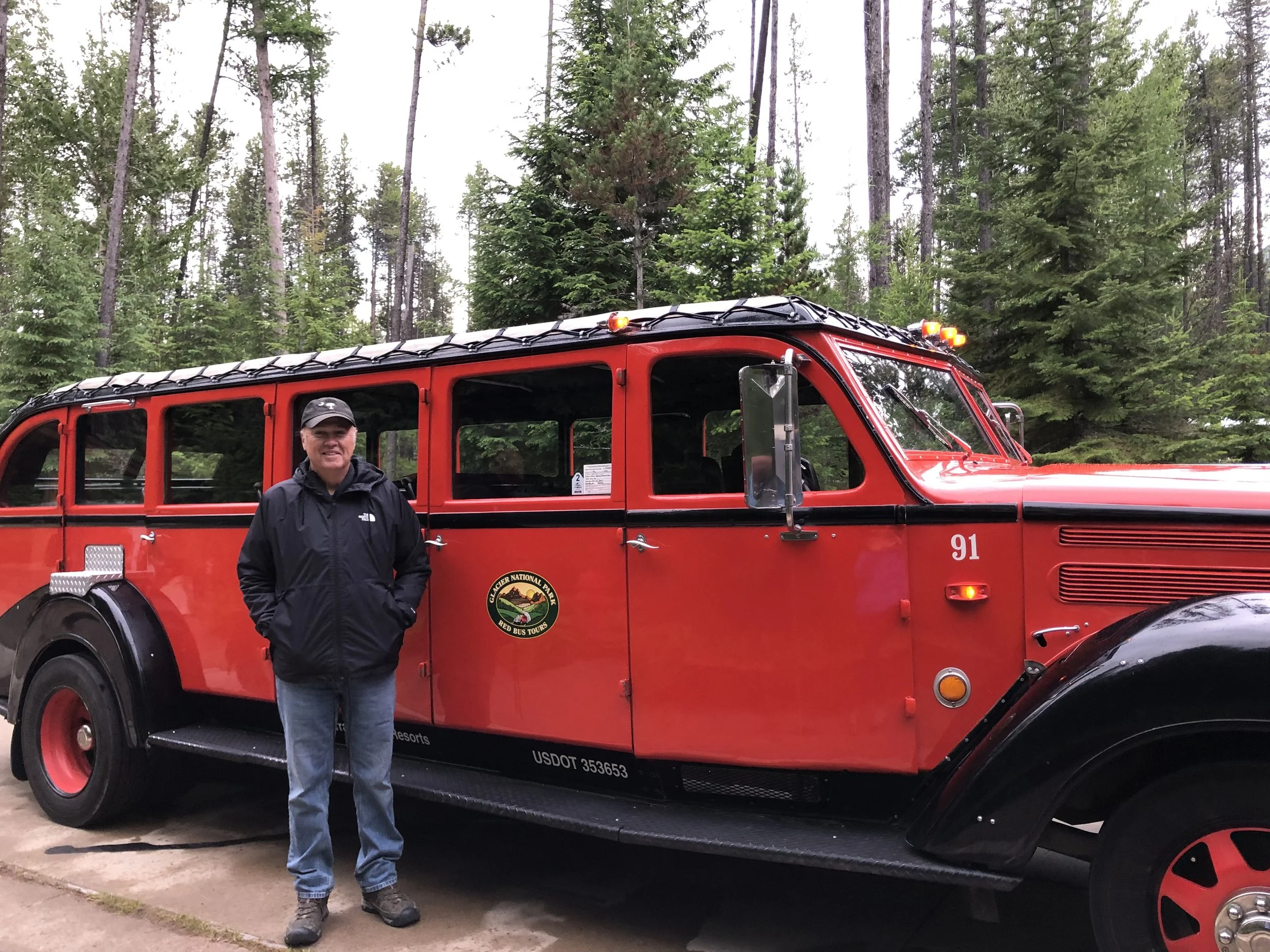 The author's dad in a black rain coat standing next to a Red Bus