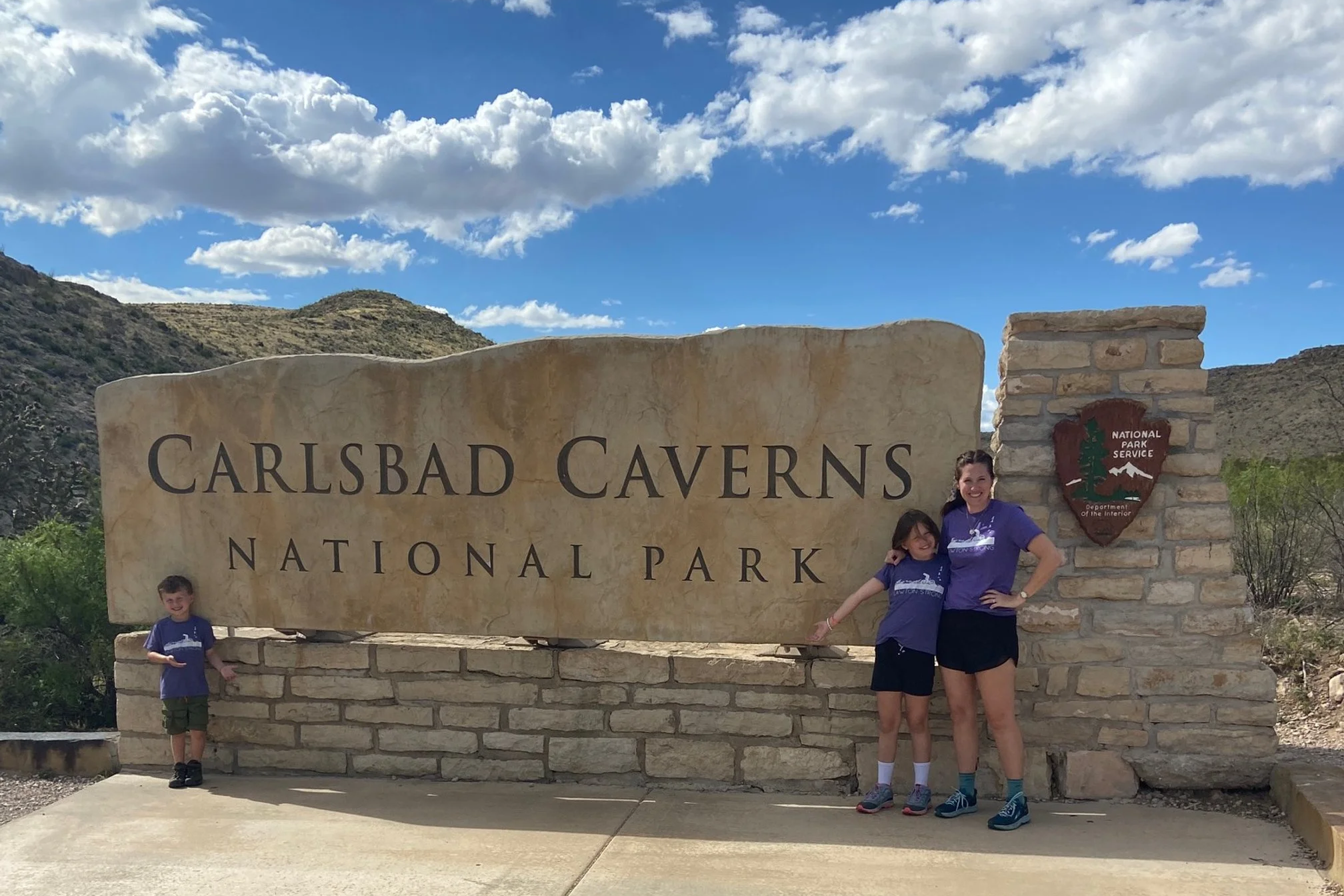 The author and her 2 children wearing purple shirts and black shorts at the entrance sign to Carlsbad Caverns National Park