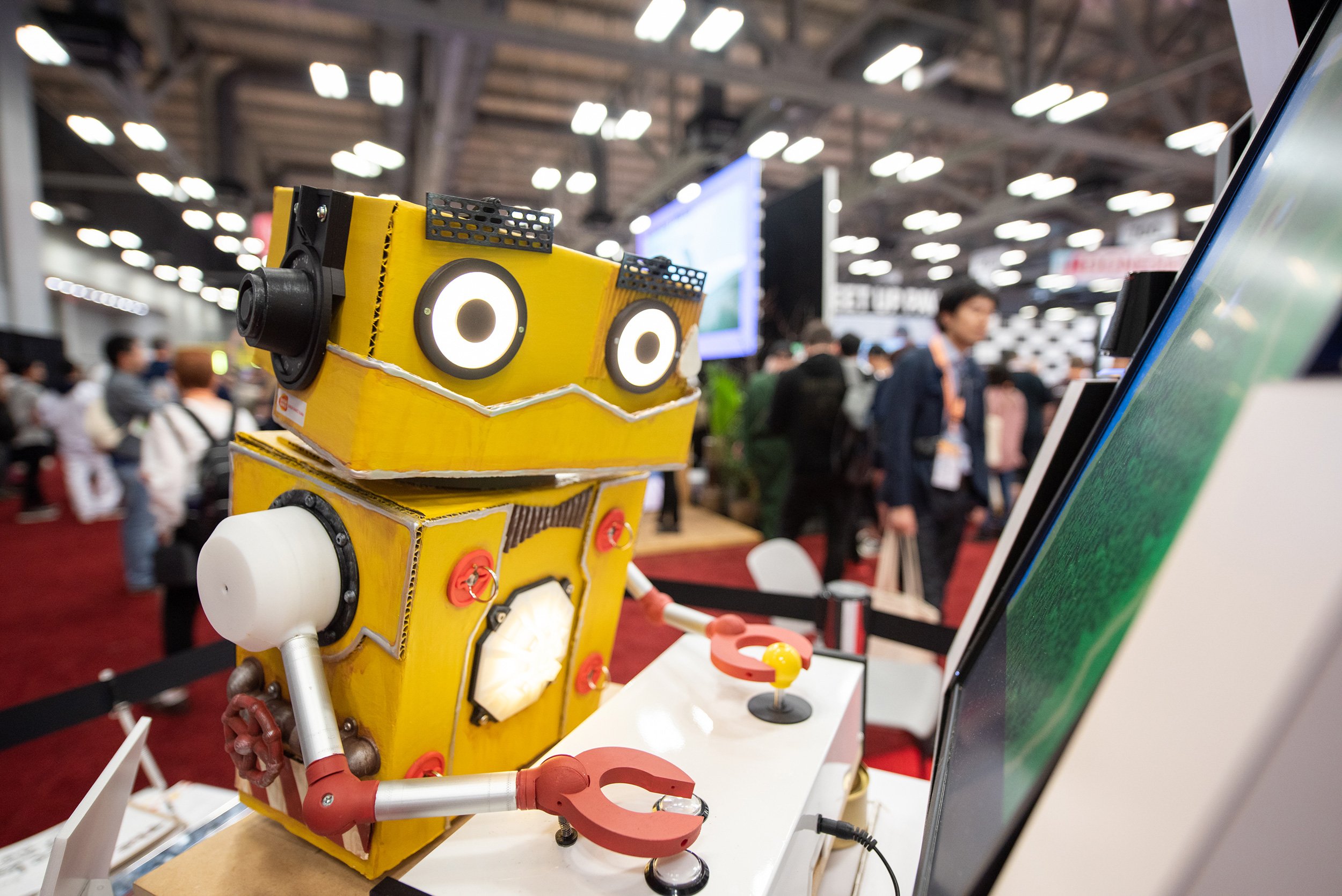 A robot made of yellow cardboard with large round eyes and a digital display on its chest, displaying a white light. The robot has a black speaker on each side of its head, and a white pipe-like arm with a clamp attached to it. It is standing at a booth with a computer monitor and a small red and yellow magnifying glass. The background shows an event with people walking around and exhibition booths with screens.