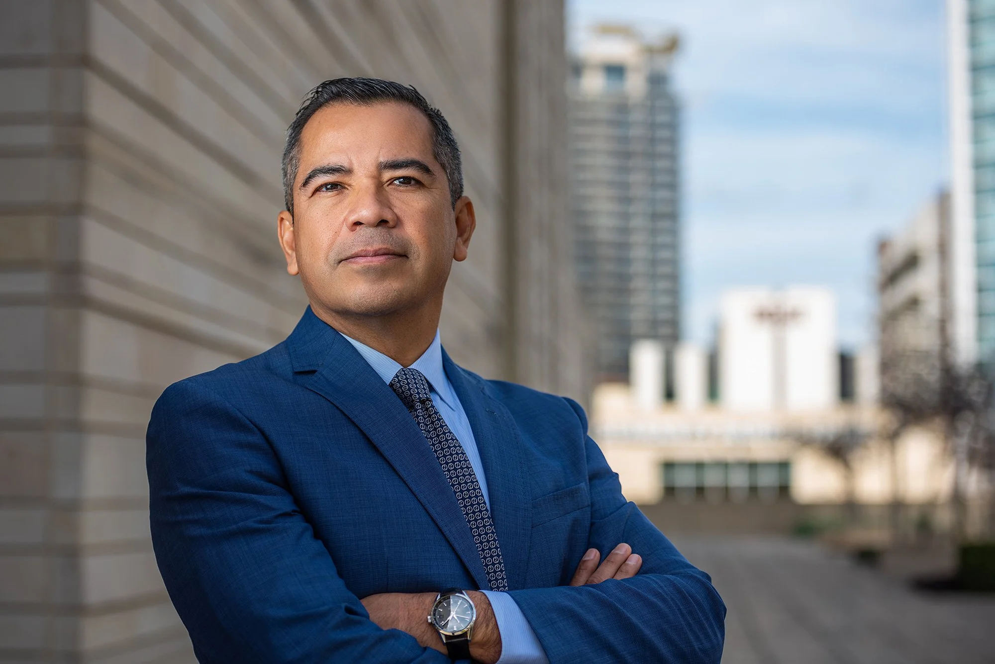 A confident man in a blue suit standing outside in an urban area with modern buildings in the background.