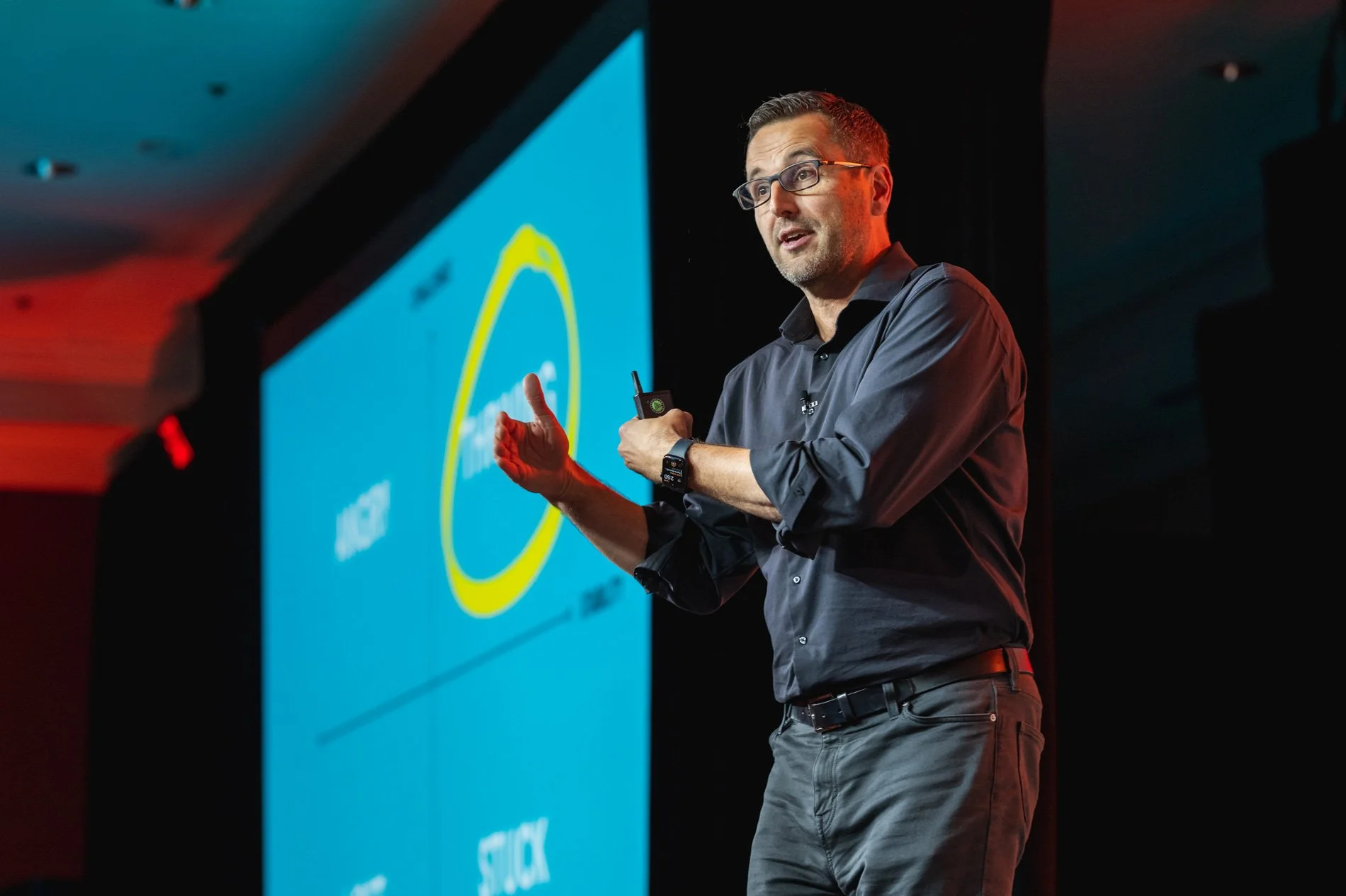 A man giving a presentation on stage with a large screen behind him showing a graphic with a yellow circle and some text.